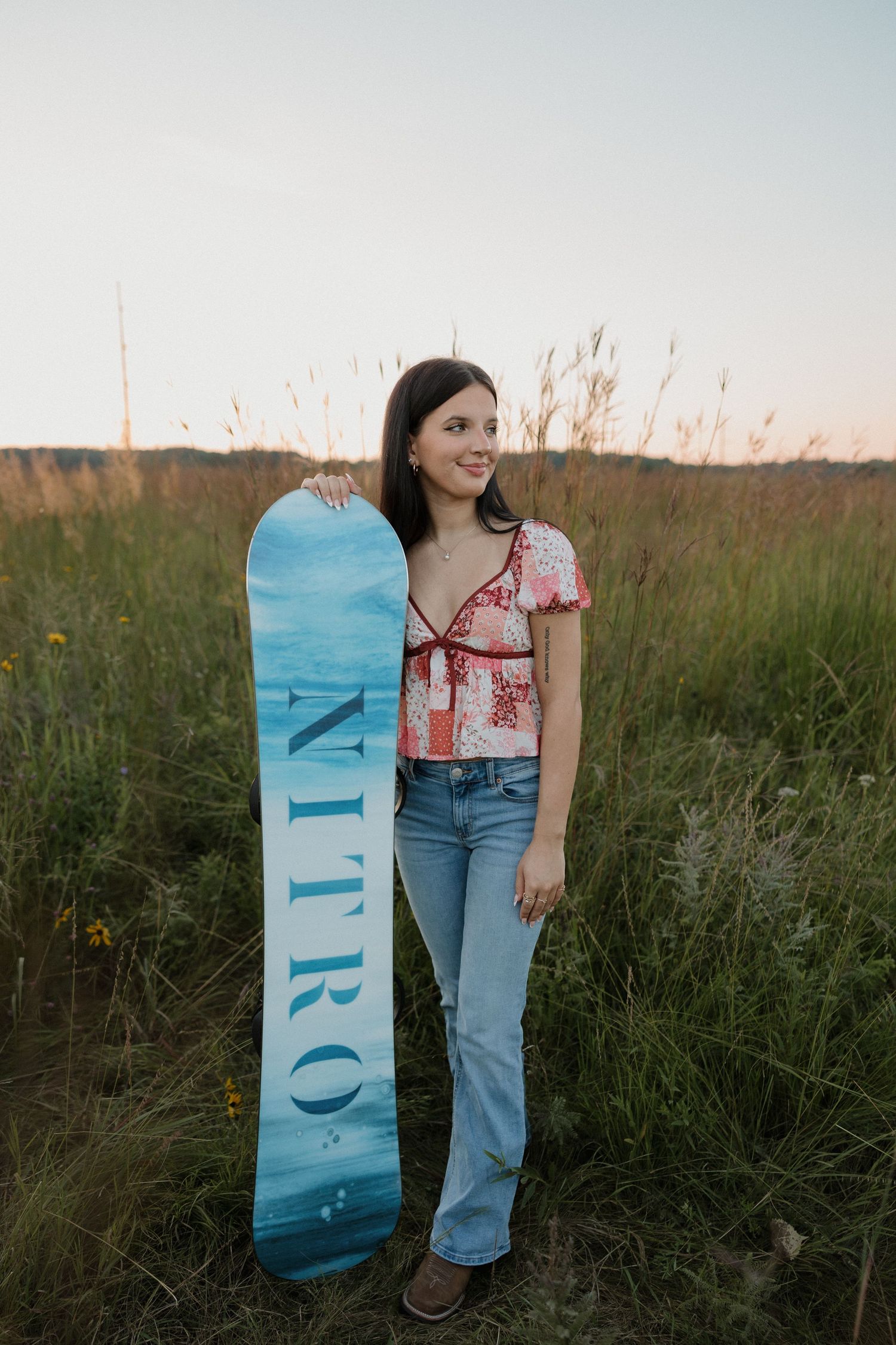 Dark-haired teen girl posing with a blue Nitro snowboard in a green field at sunset.