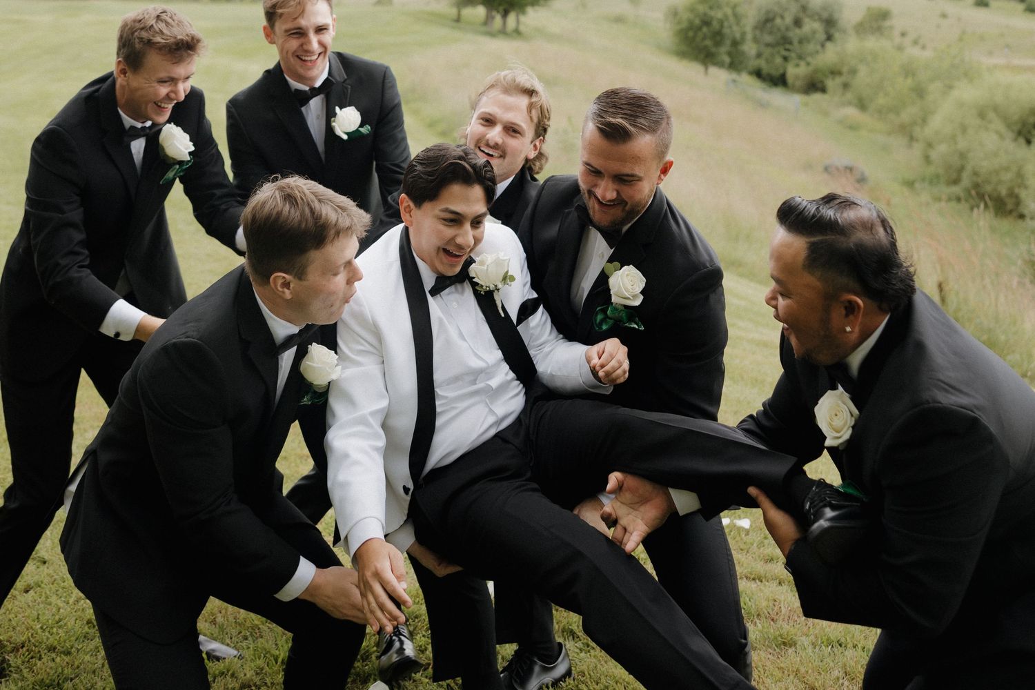 Groomsmen in black suits and tuxedos pose playfully on a grassy hillside during wedding photos.