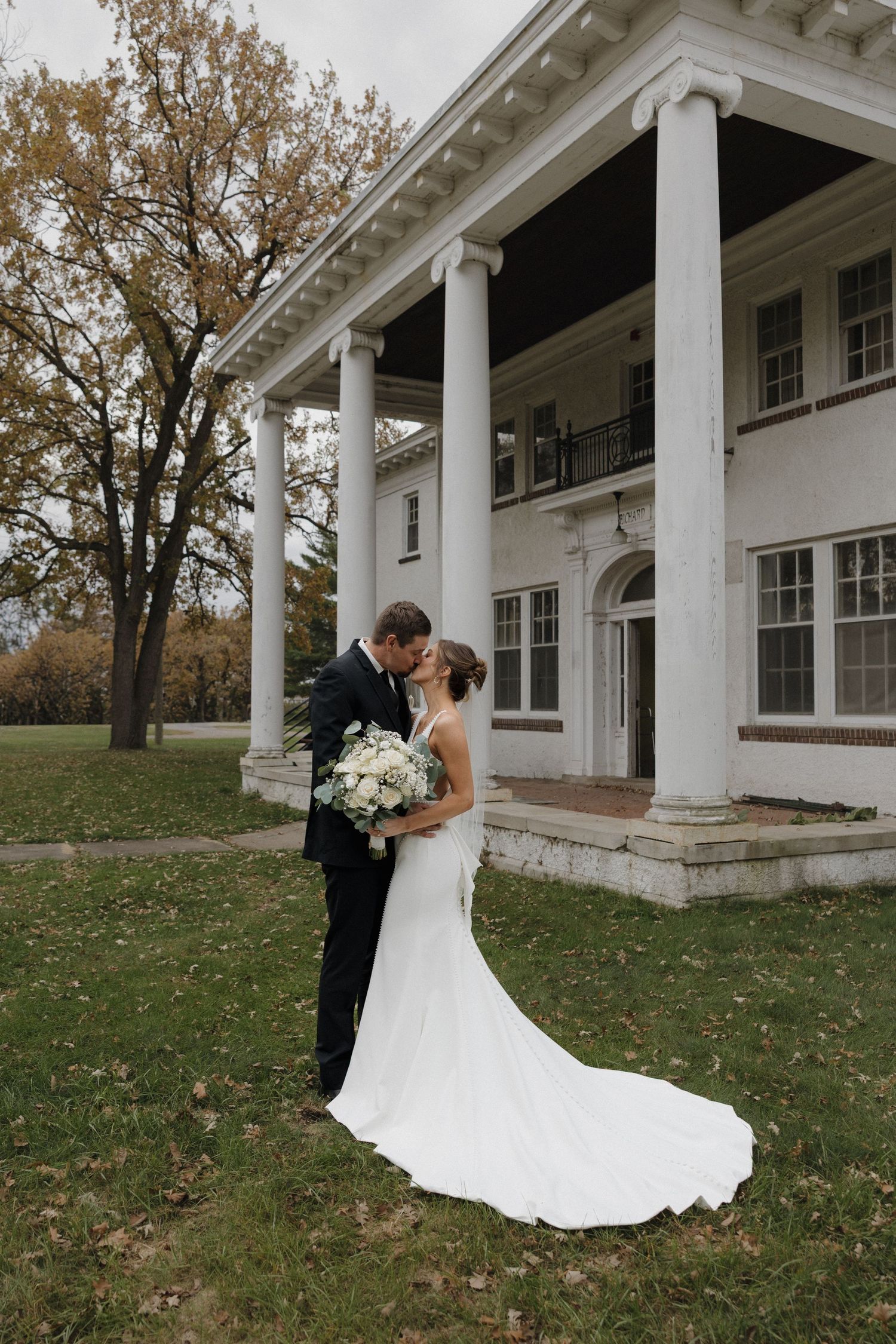 Bride and groom kissing in front of a white colonial mansion surrounded by autumn trees.