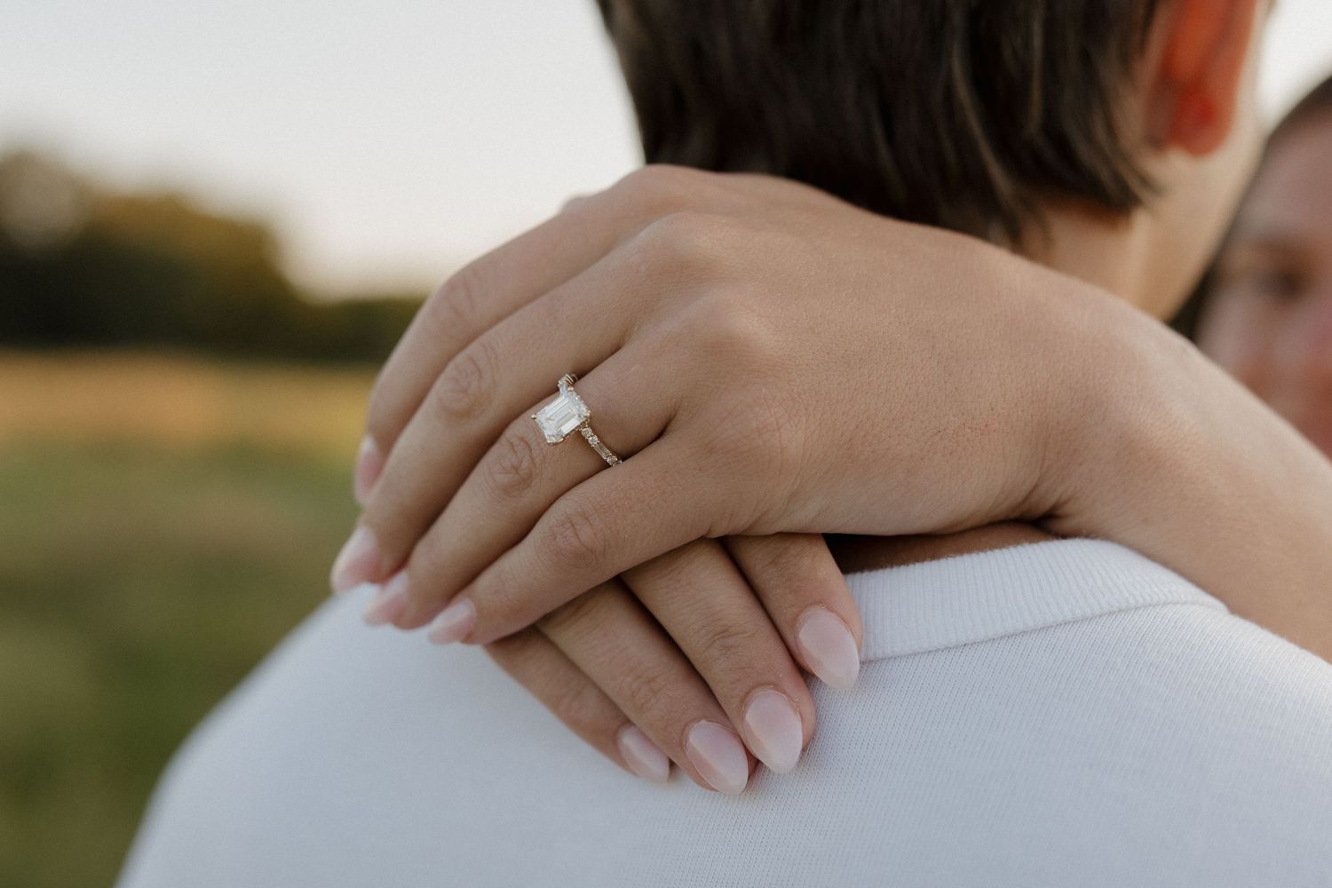 Close-up of a woman's hand showing a diamond engagement ring resting on her partner's shoulder.