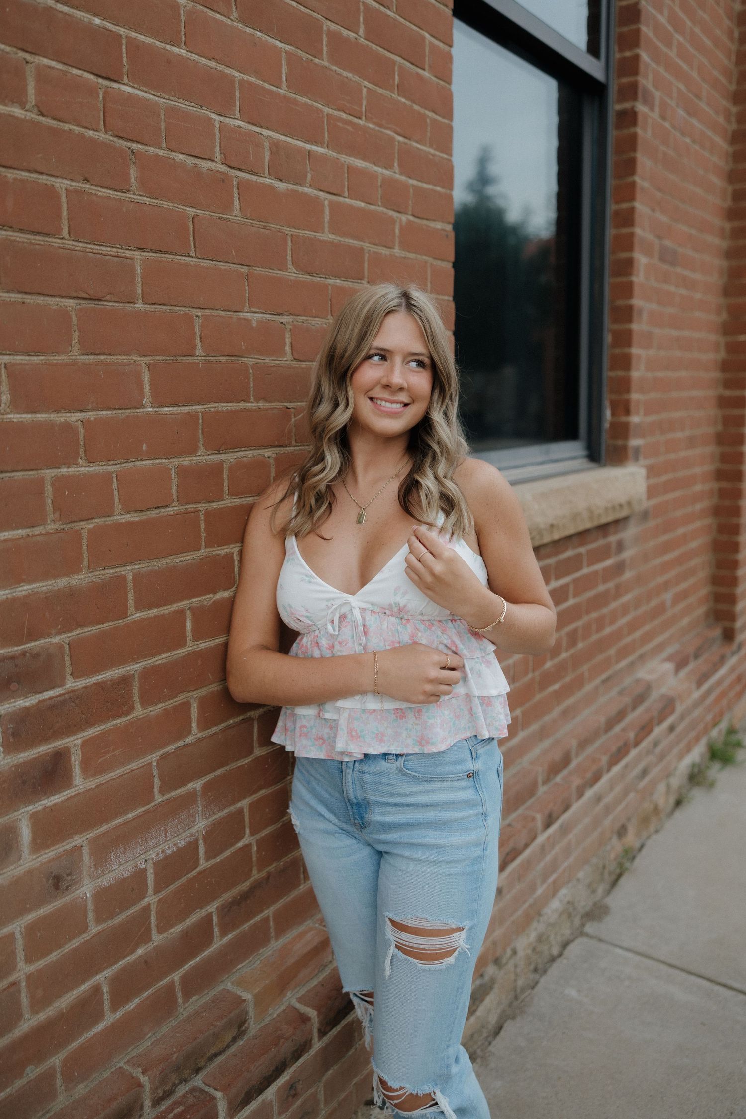 Smiling blonde woman in floral cami top and ripped jeans leans against a red brick wall.