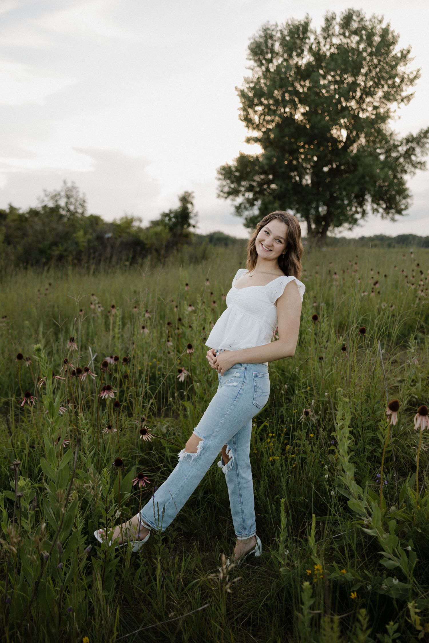 Smiling young woman in a white top and ripped jeans standing in a lush wildflower meadow.