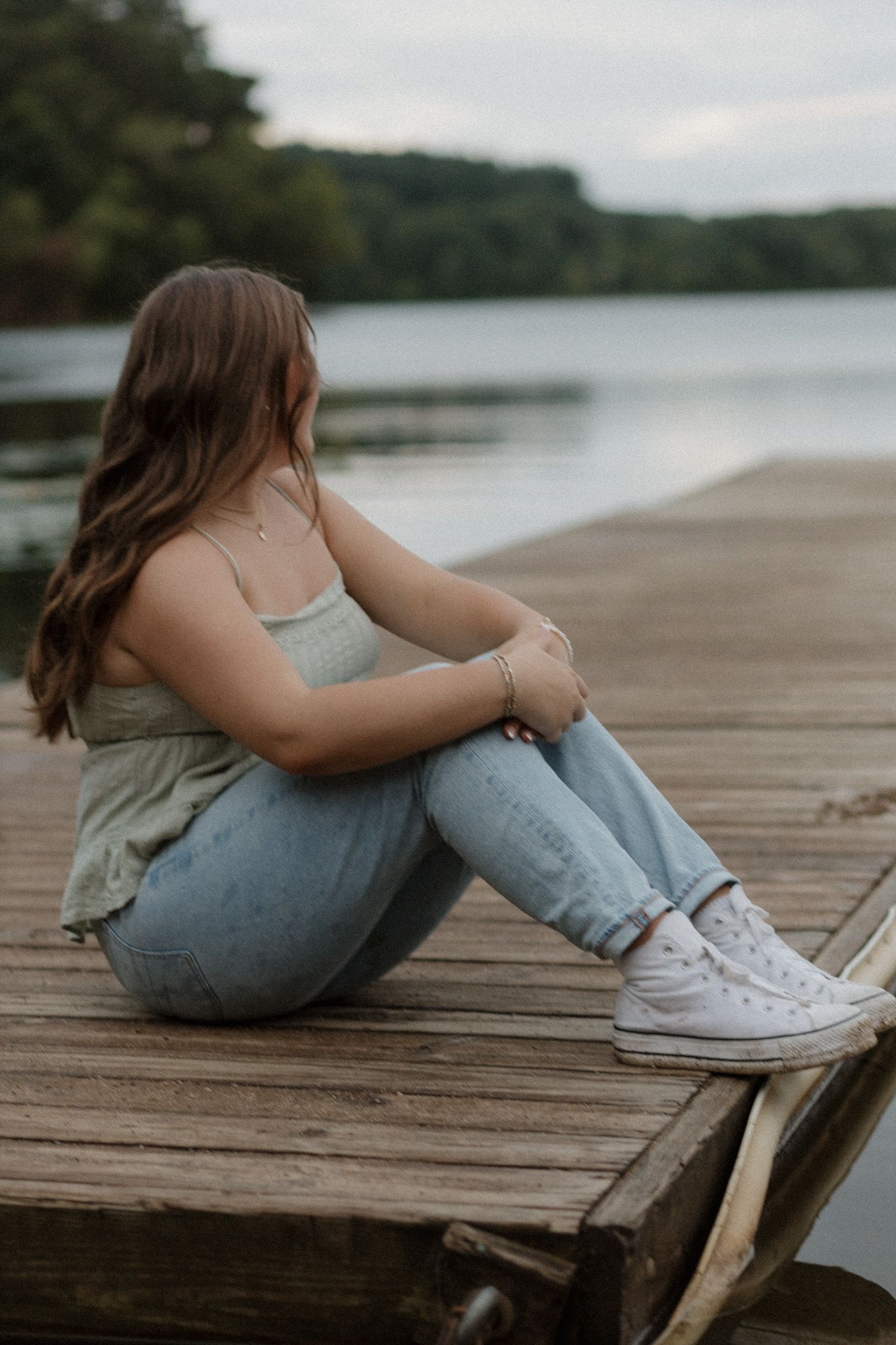 Teen girl in green top and jeans sitting on wooden dock gazing at serene lake.