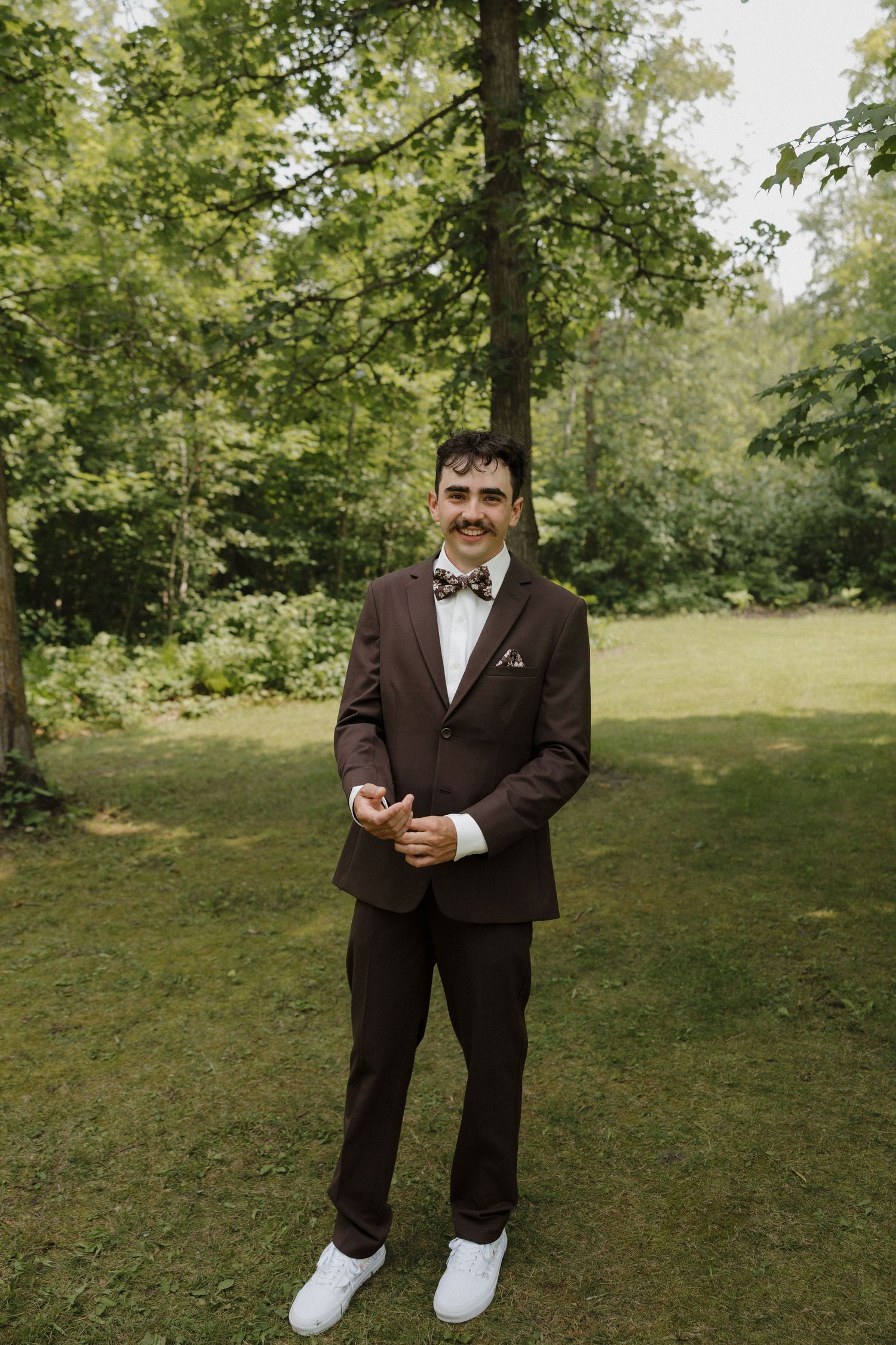 Person in a dark suit and bow tie checking their phone while standing in a lush green outdoor setting.