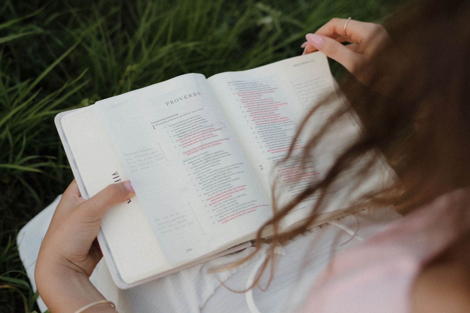 Young woman reading open book while sitting in green grass outdoors.