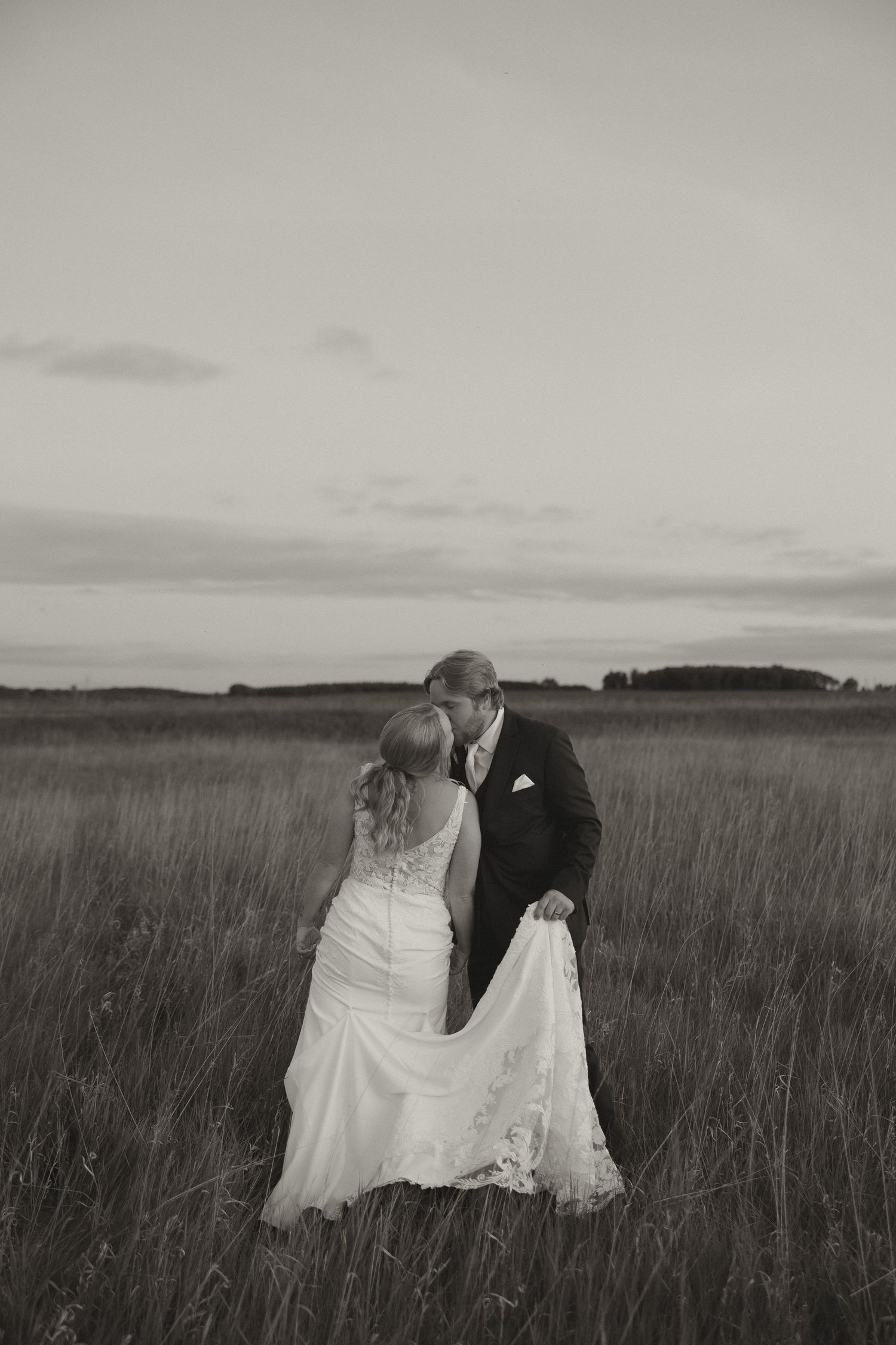 Black and white romantic photo of bride and groom kissing in an open field at dusk.