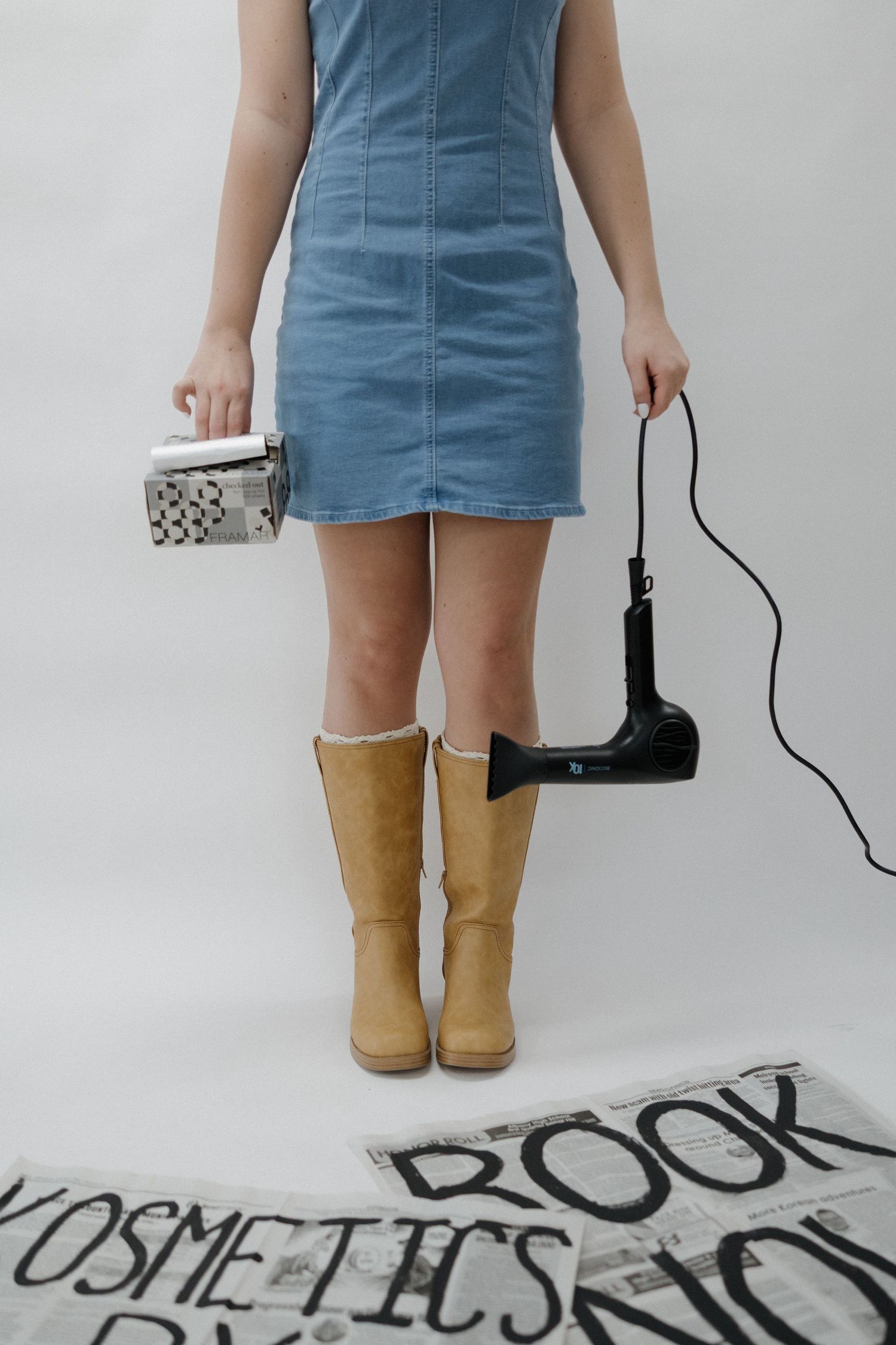 Woman in blue denim dress and tan boots holding a hair dryer and styling tool over newspaper signs.
