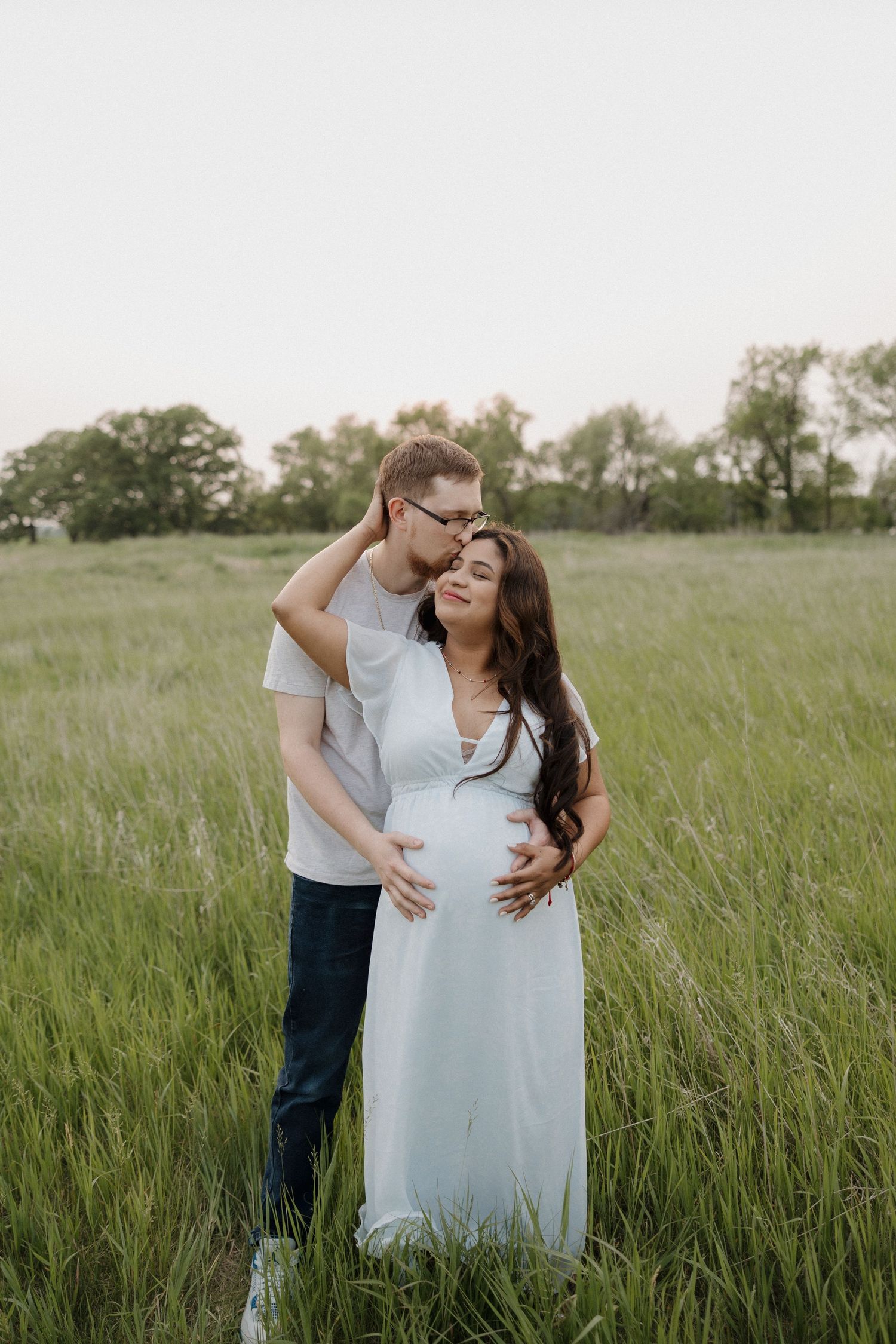 Couple embracing in green field, man kissing pregnant womans head while holding her bump.
