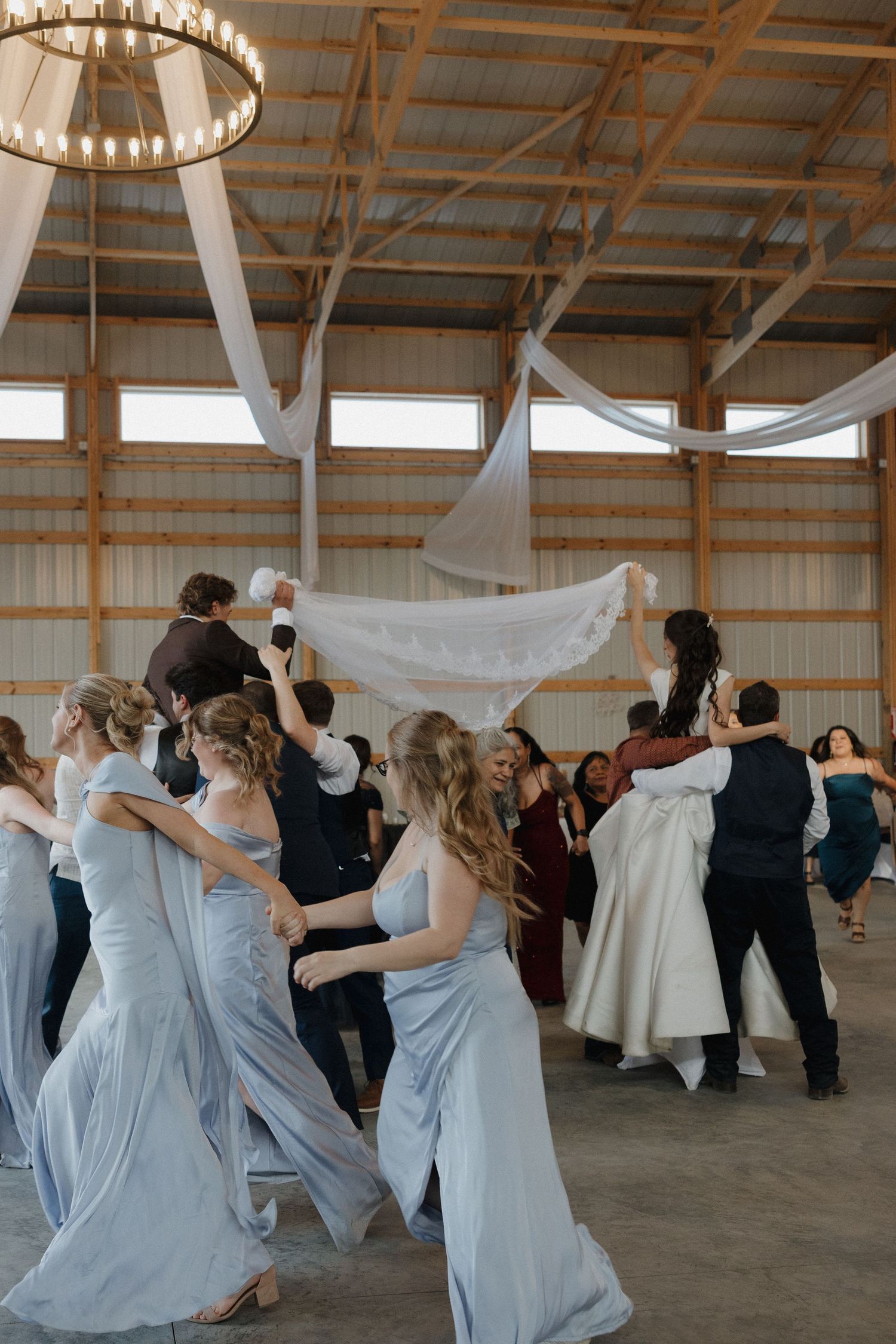 Wedding party celebrates on dance floor in rustic barn venue with light blue bridesmaid dresses and white fabric decor.