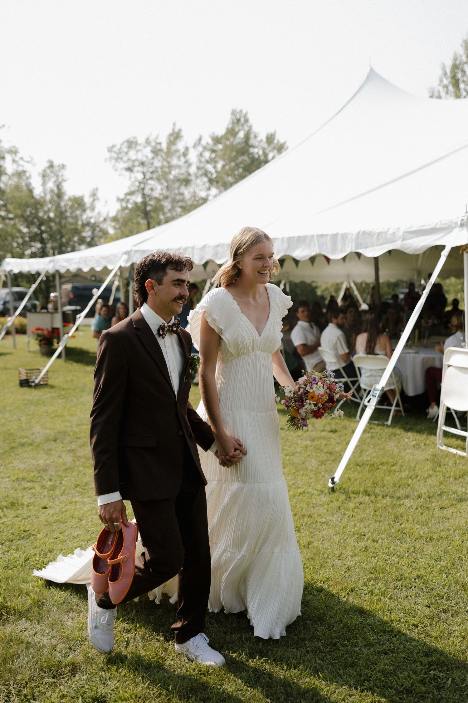 Newlyweds walking hand-in-hand across a sunny lawn with a white reception tent in the background.