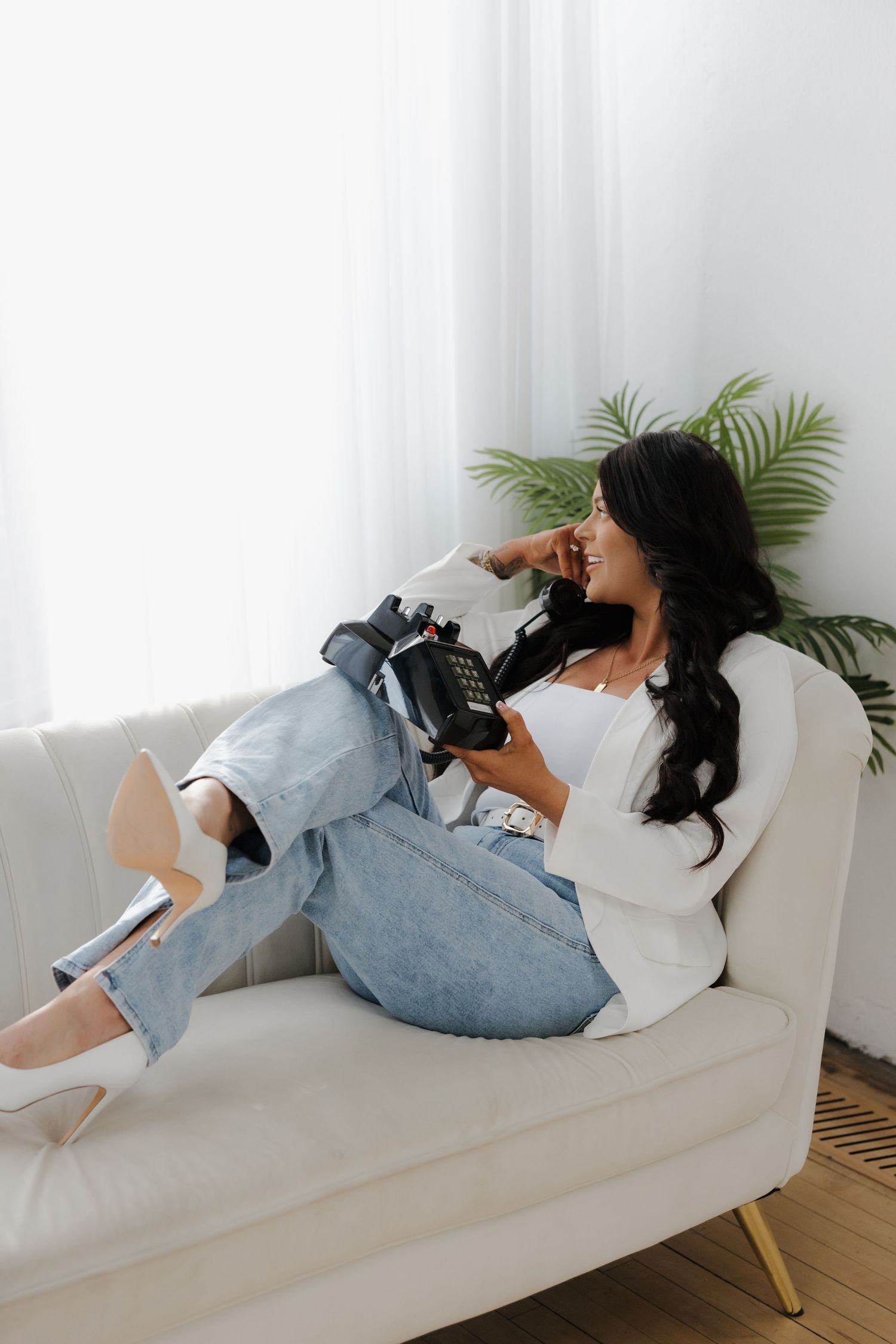 Smiling brunette woman relaxing on white sofa holding vintage telephone by window.