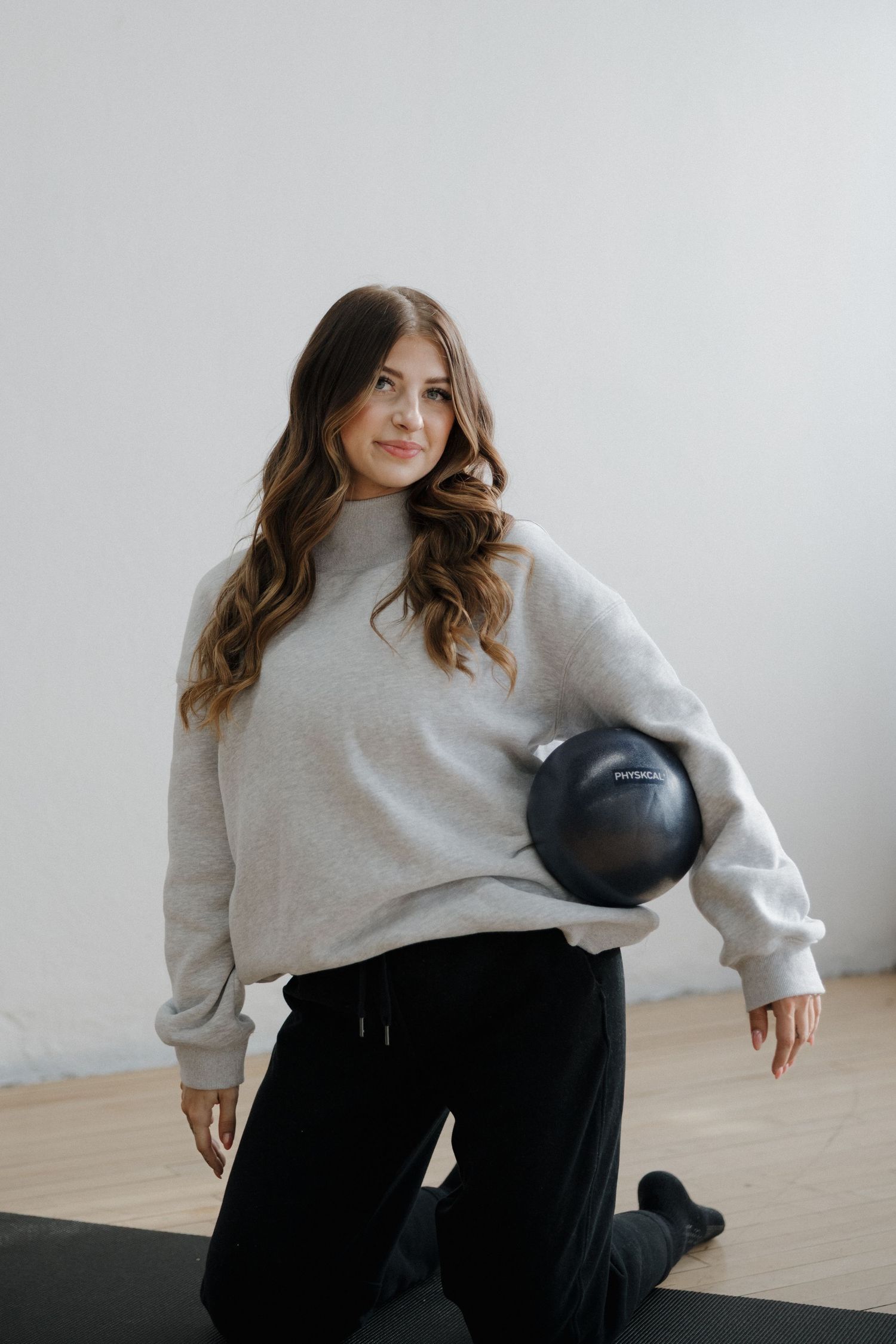 Woman in gray sweatshirt and black pants holding a dark pilates ball on a yoga mat.