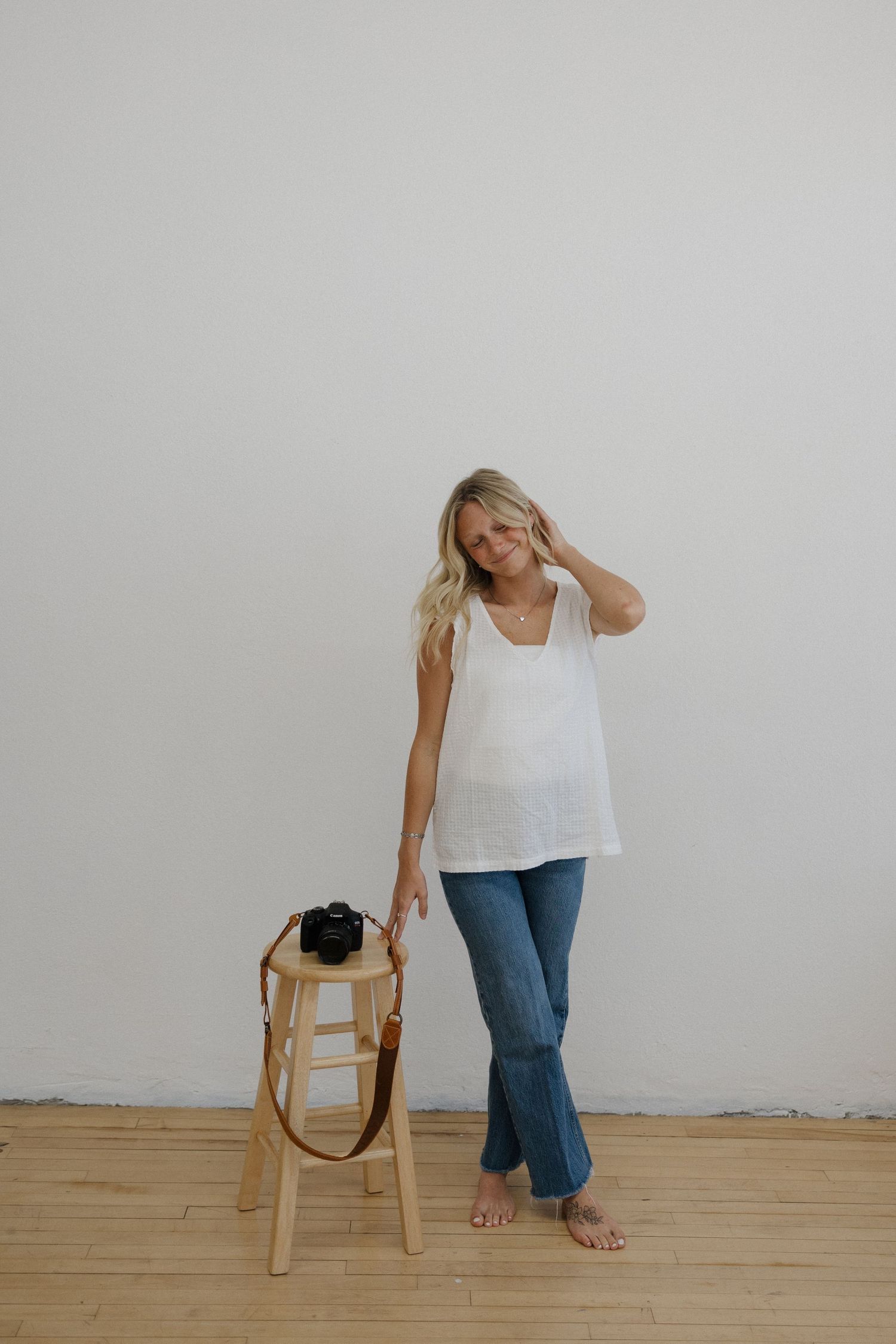 Smiling blonde woman in white tank top standing beside a wooden stool with a camera.