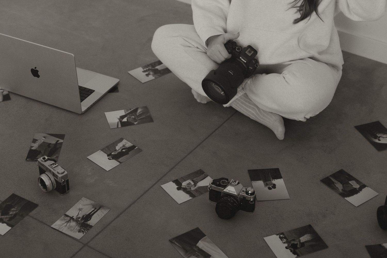 Photographer sitting on floor surrounded by printed photos and cameras, reviewing work with laptop nearby.
