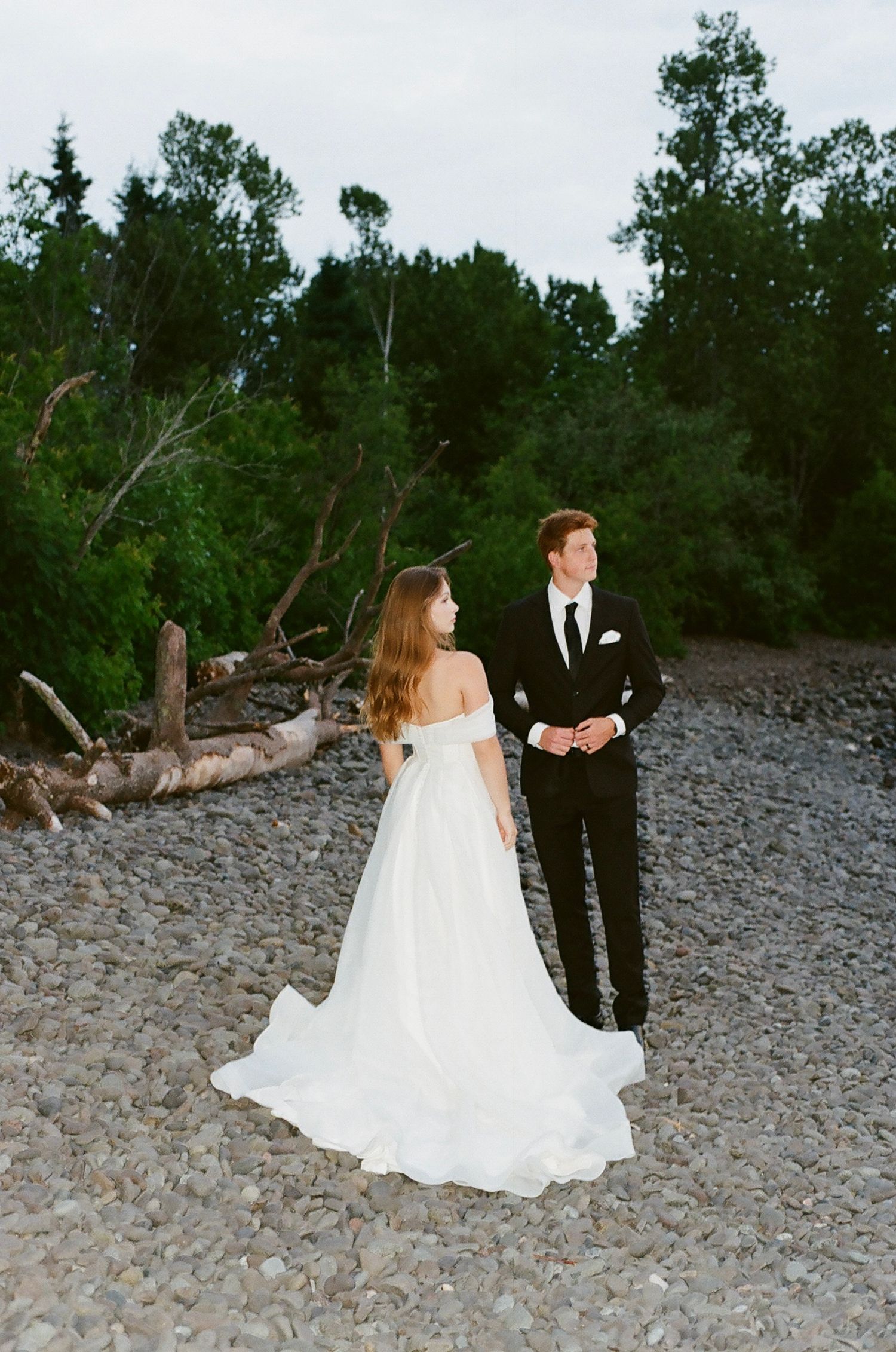 Bride in off-shoulder white gown and groom in black tuxedo stand on rocky shore with driftwood and trees behind them.