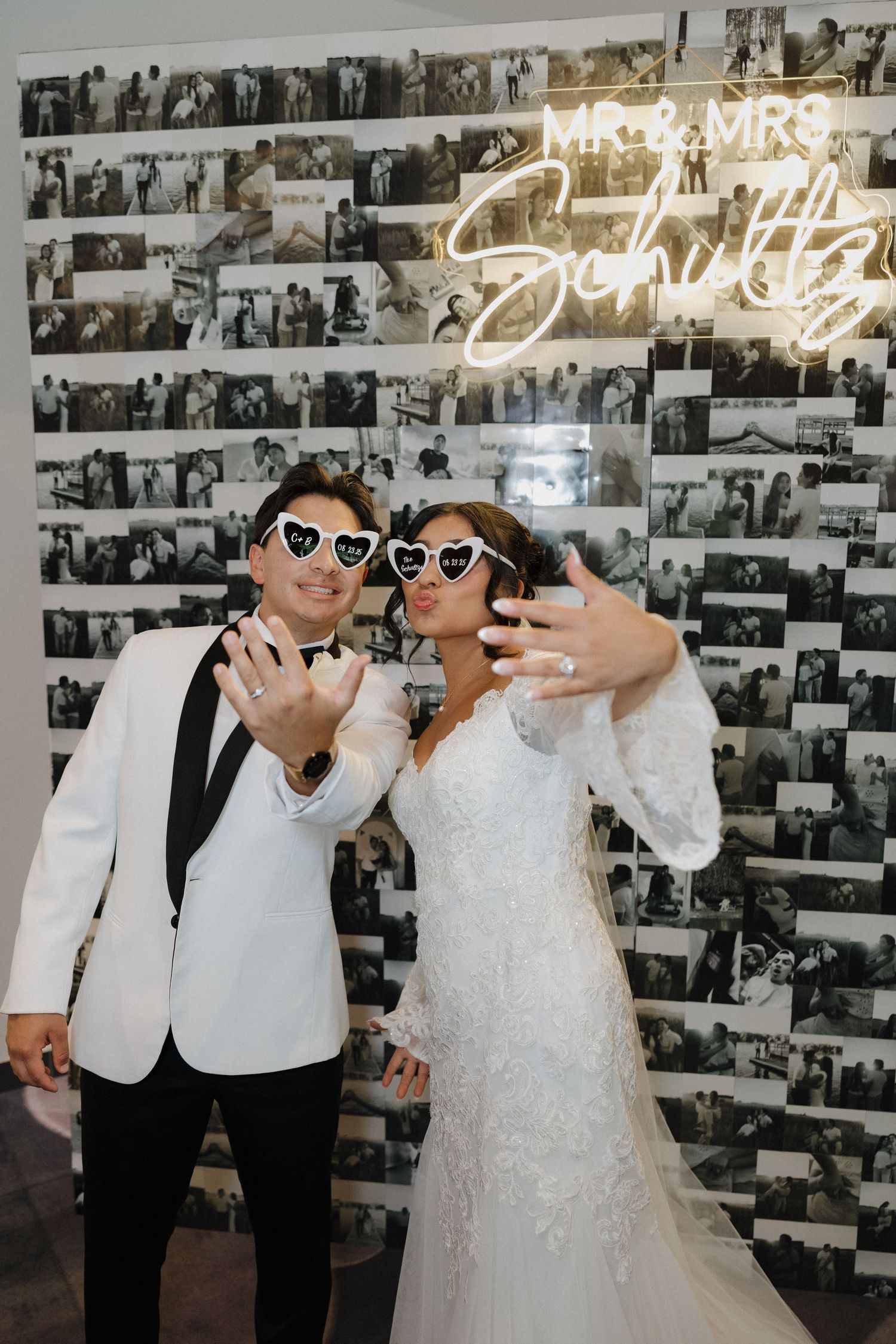 Wedding couple wearing heart-shaped sunglasses posing in front of photo wall with neon sign.
