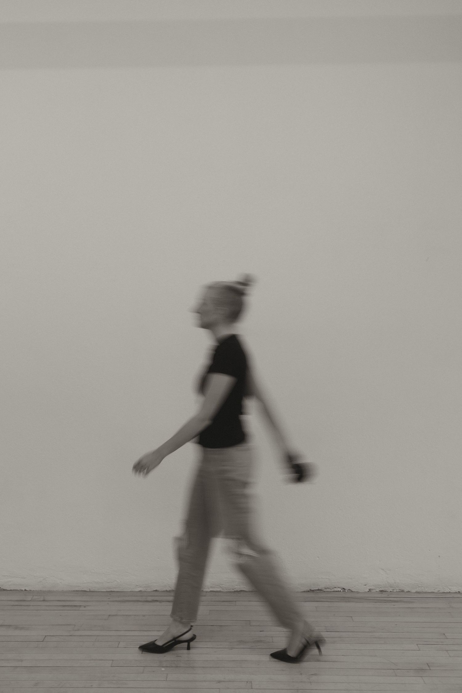 Blurred motion black and white photo of woman walking in heels against white wall.