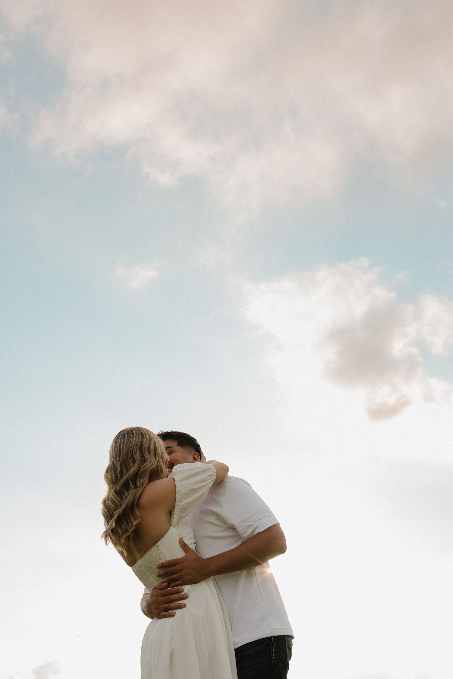 Romantic couple embracing and kissing against a beautiful blue cloudy sky backdrop outdoors.