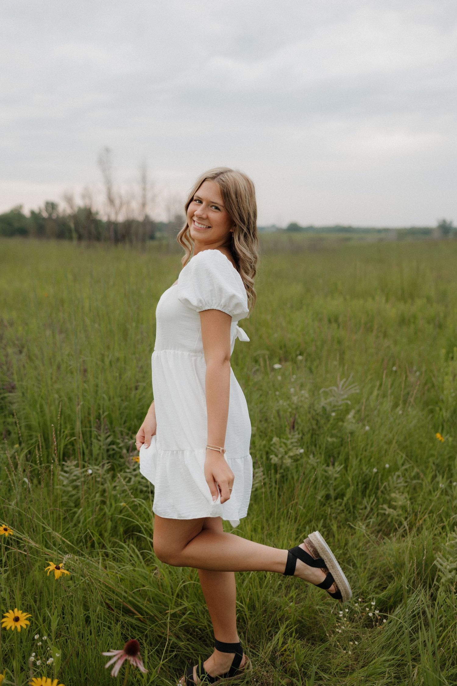 Smiling blonde woman in white puff-sleeve dress poses playfully in a green grassy field.