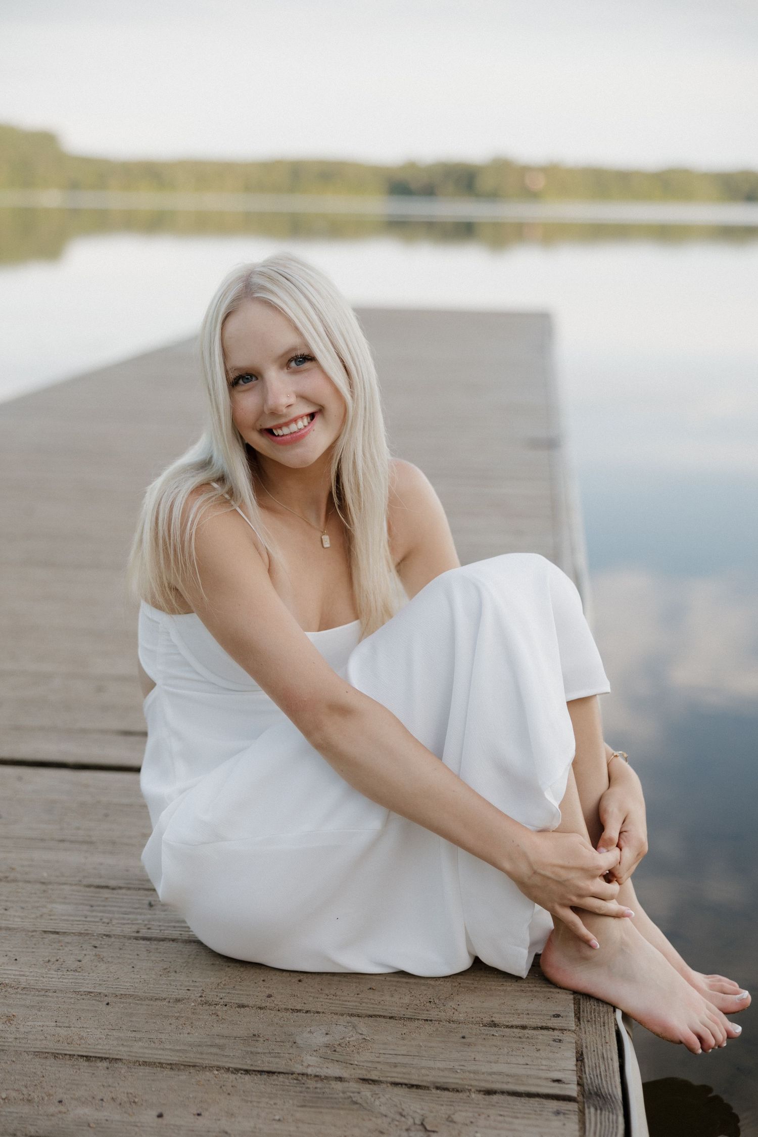 Smiling blonde woman in white outfit sitting barefoot on a wooden dock by a serene lake.