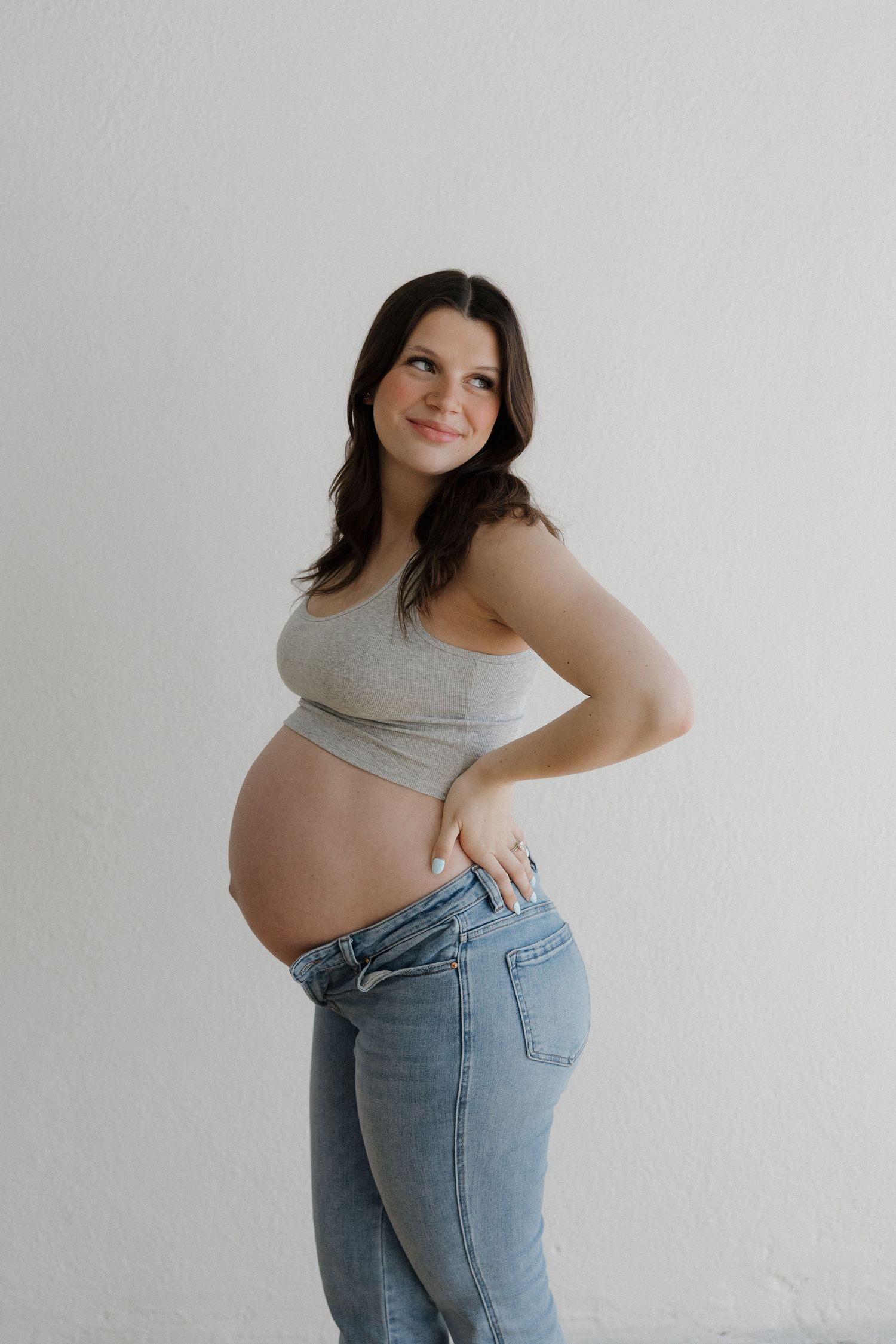 Smiling brunette pregnant woman in gray crop top and jeans posing against white wall.