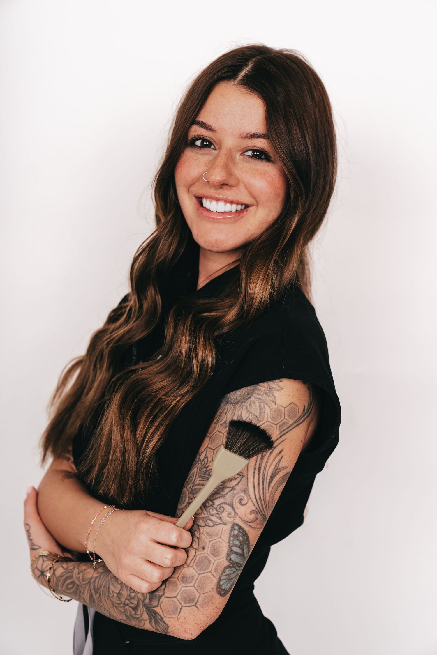 Smiling woman with long brown hair, sleeve tattoos, holding a makeup brush, wearing a black top against white background.
