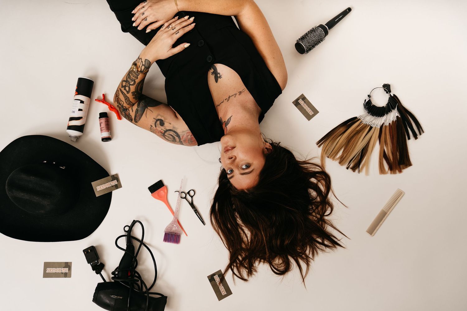Tattooed woman lying on white floor surrounded by hair tools, extensions, scissors, and beauty products.