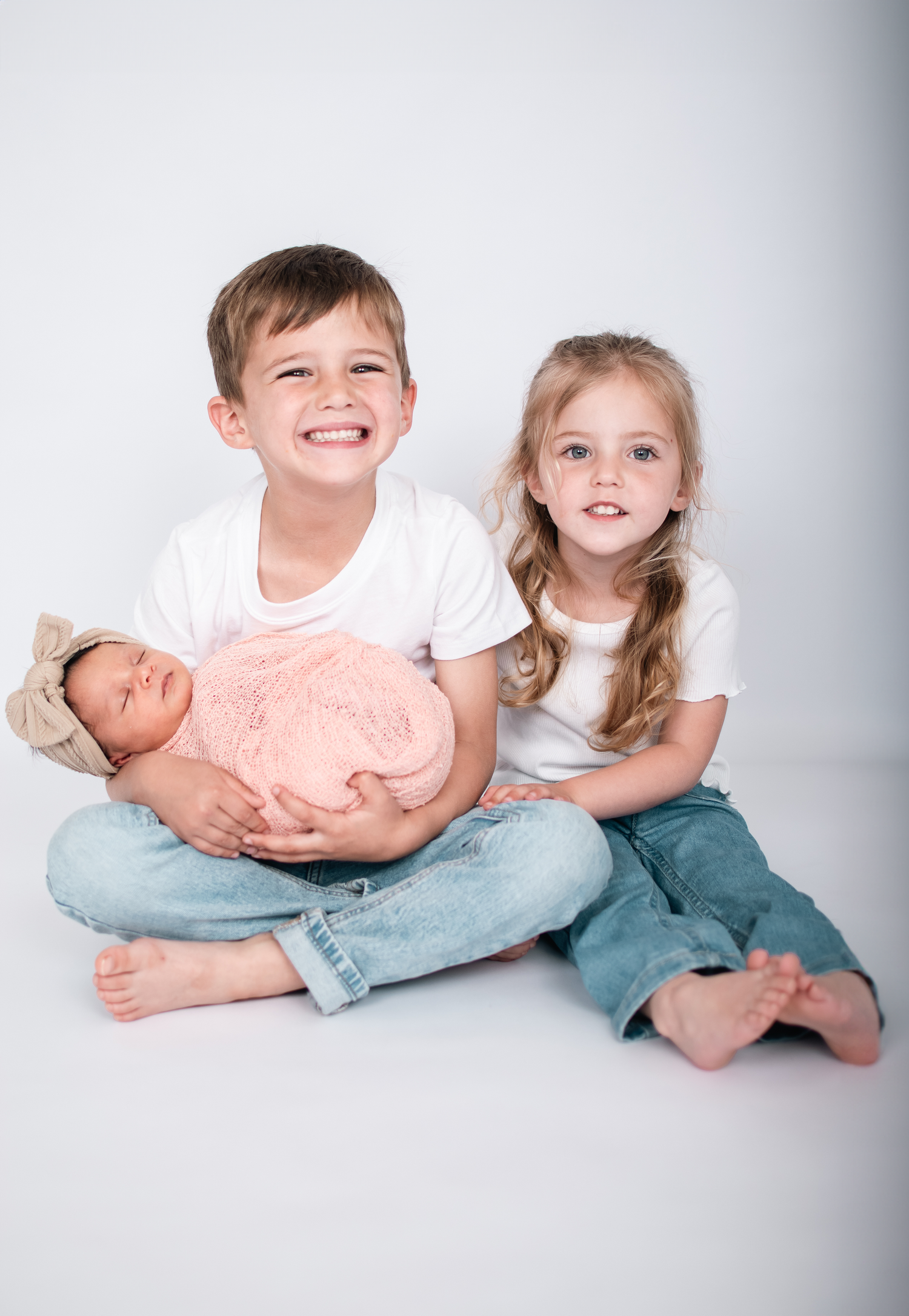 Studio portrait of siblings sitting together with newborn baby wrapped in pink.