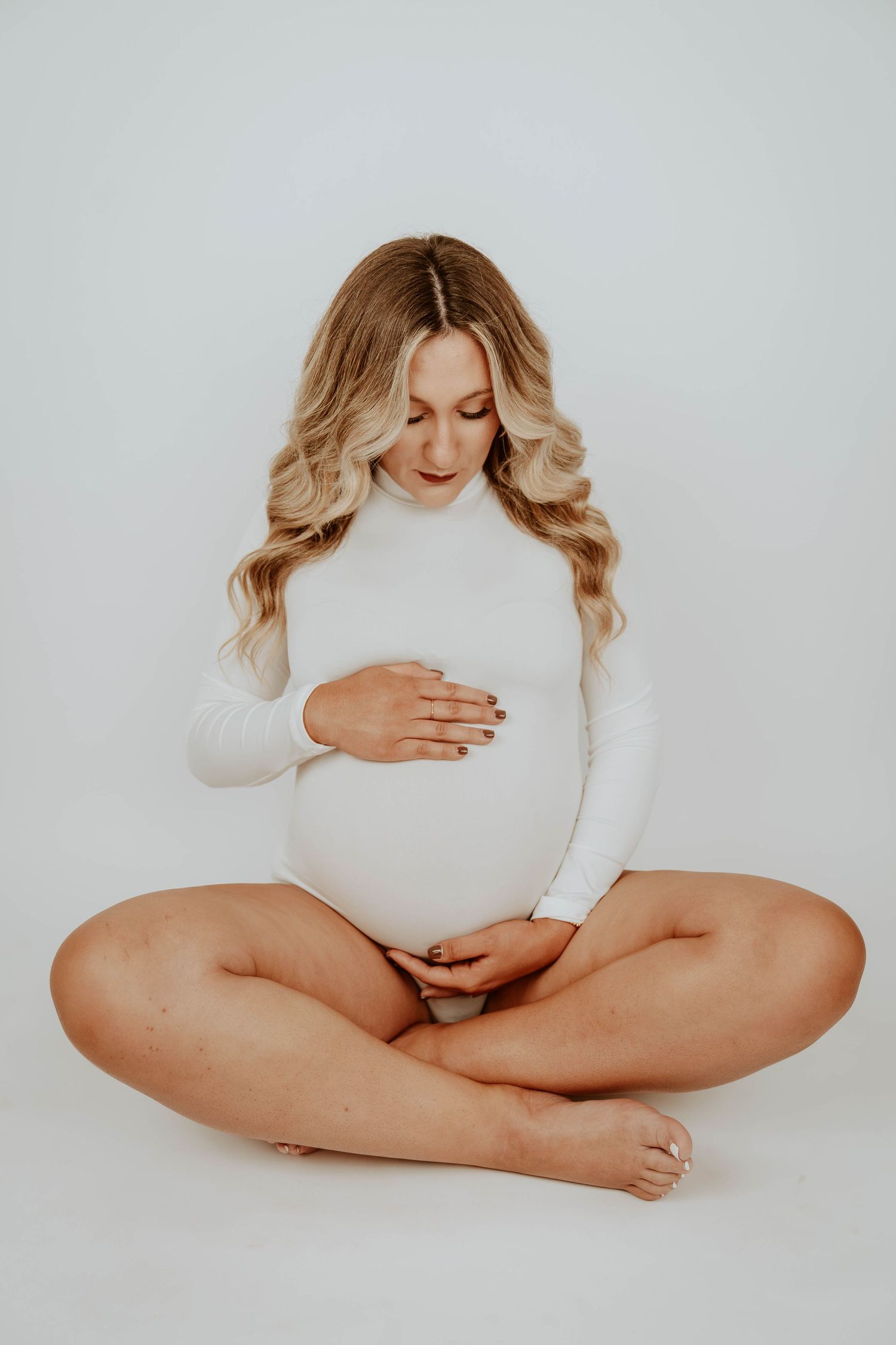 A pregnant woman in white sits cross-legged during a studio photoshoot.
