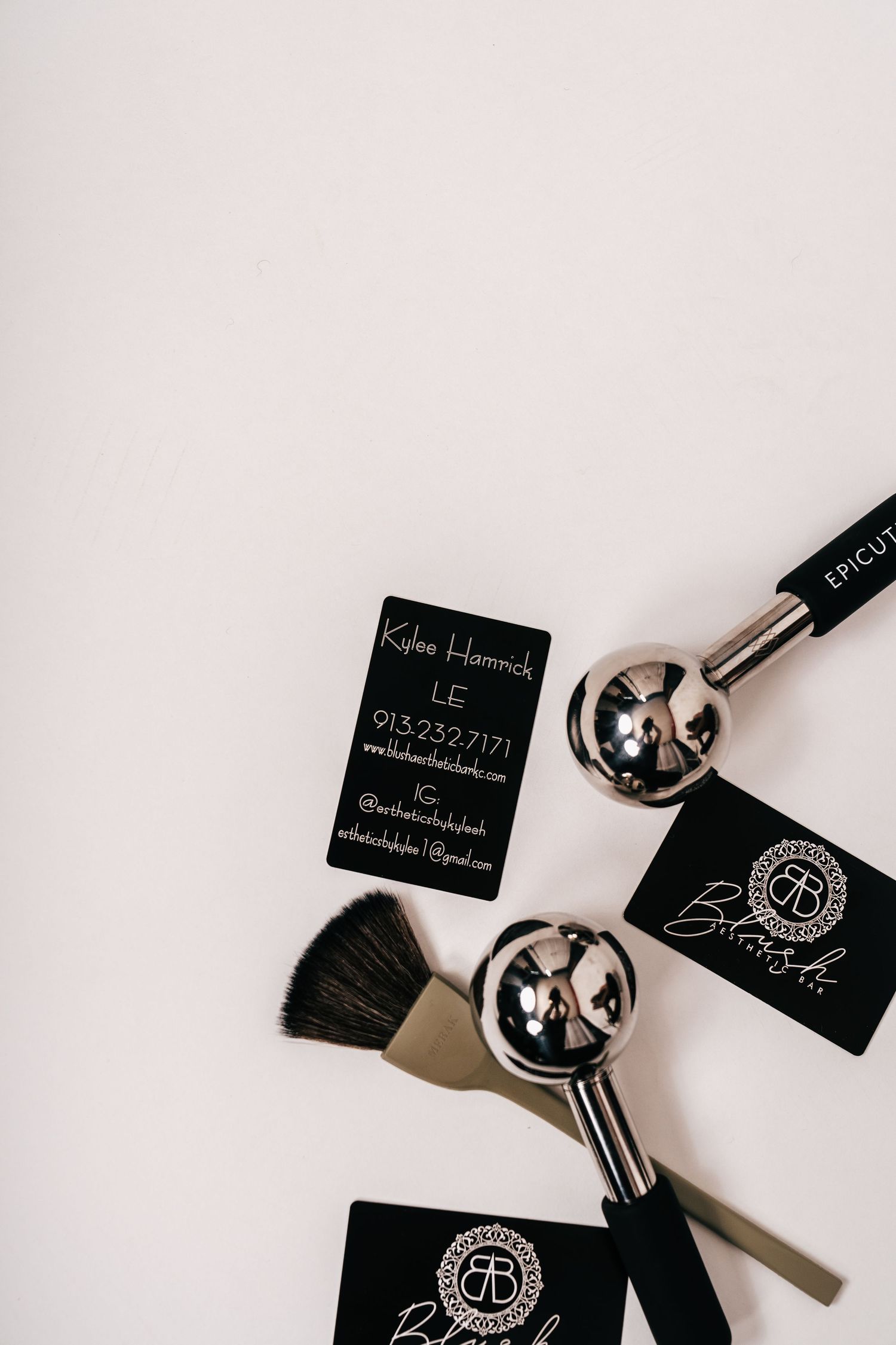 Black business cards, makeup brushes, and chrome tools arranged on white background for beauty professional Kylee Hamrich.