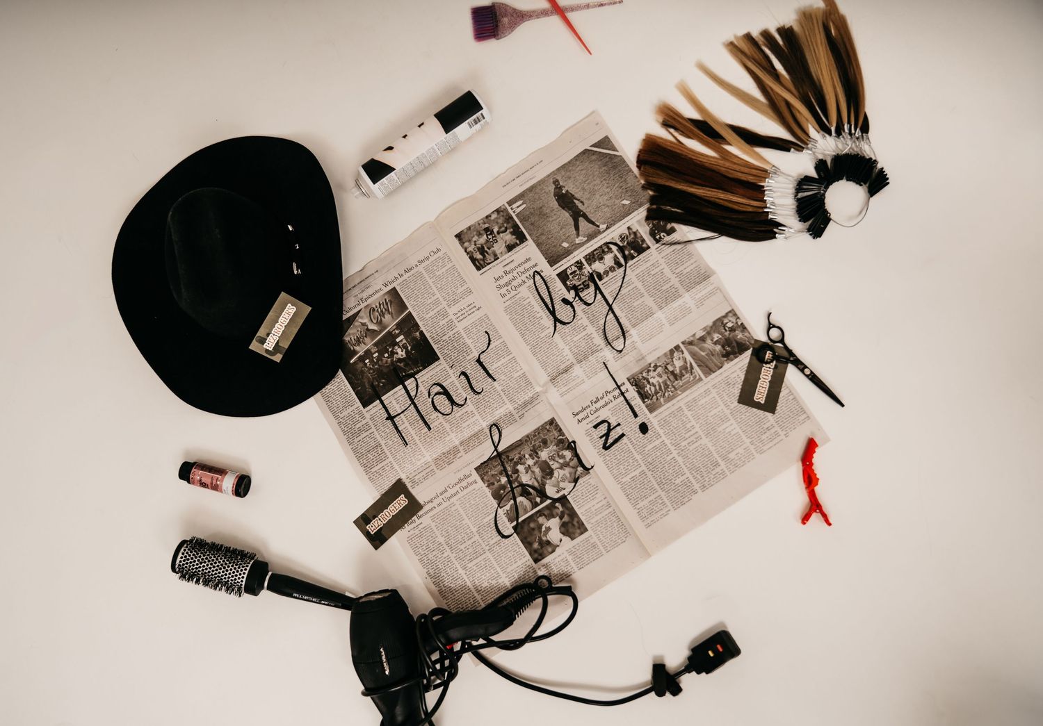 Hair styling tools, hat, extensions, and newspaper spread on white background for hair salon branding.