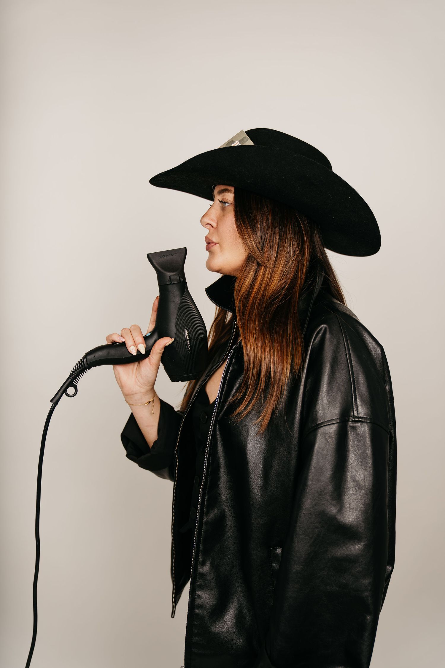 Woman in black cowboy hat and leather jacket holds hair dryer like microphone against light gray background.