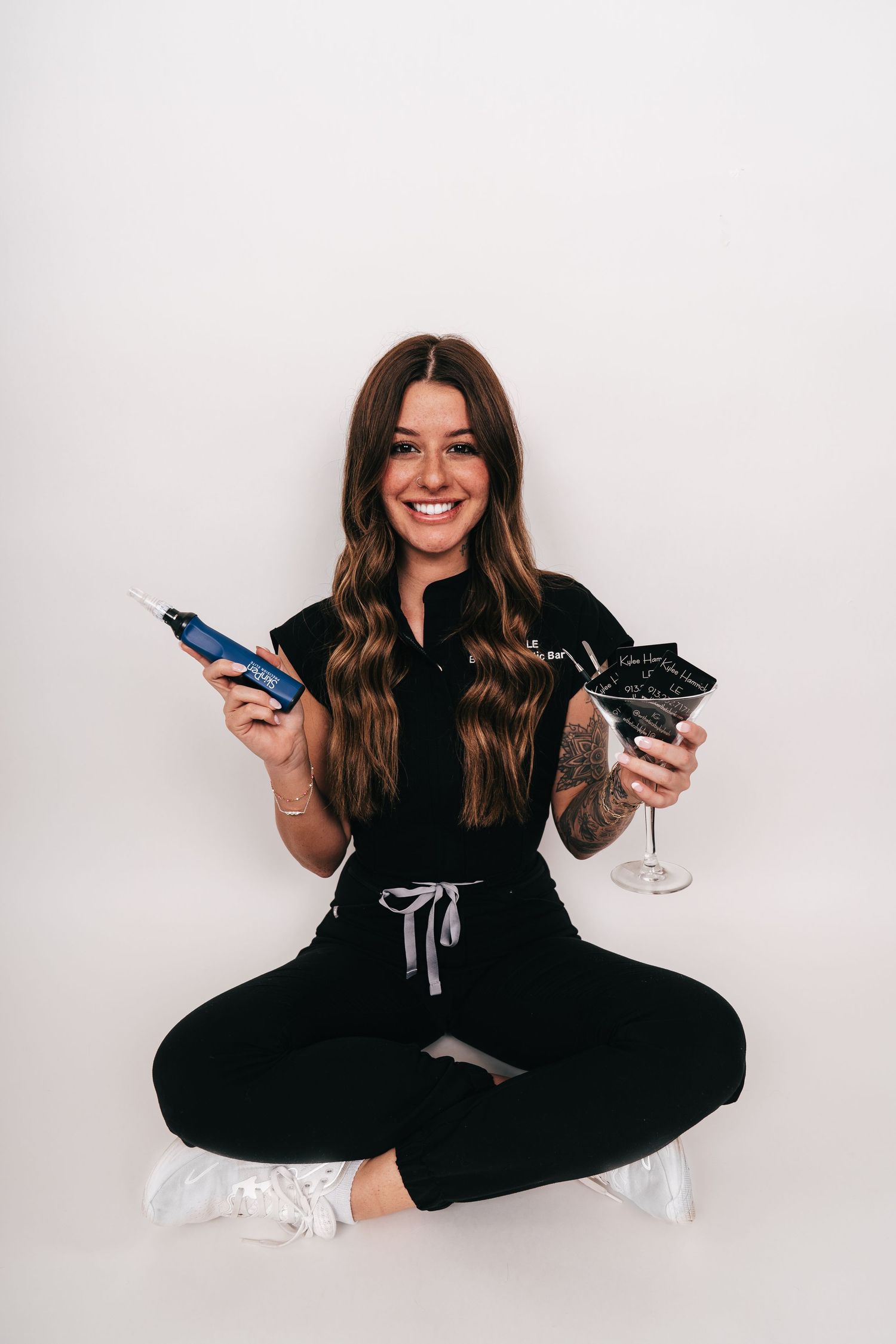 Smiling woman in black outfit sitting cross-legged, holding a blue pen and cocktail glass against white background.