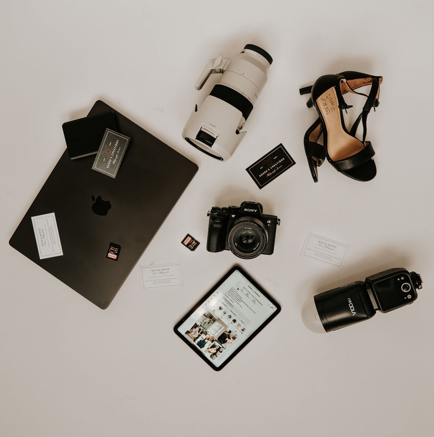 Flat lay of photography gear including a Sony camera, MacBook, tablet, flash, lens, SD card, and heels on white background.