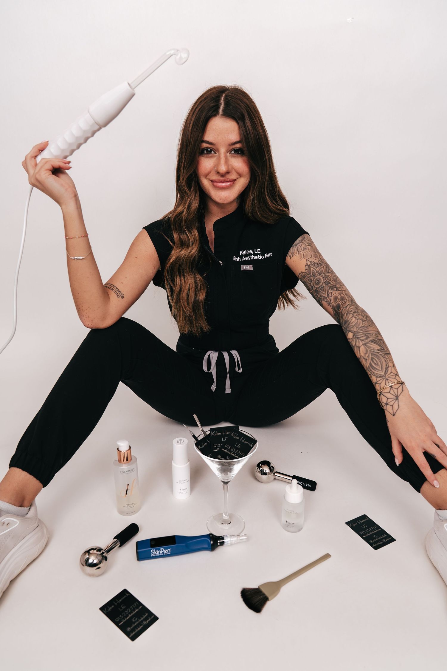 Woman in black outfit holding curling wand, sitting with beauty tools and skincare products on white background.