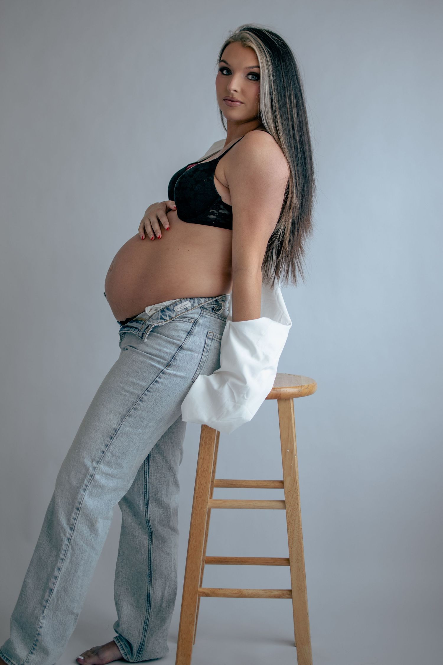 Artistic maternity photoshoot showing a woman in jeans and white top posing against grey backdrop.