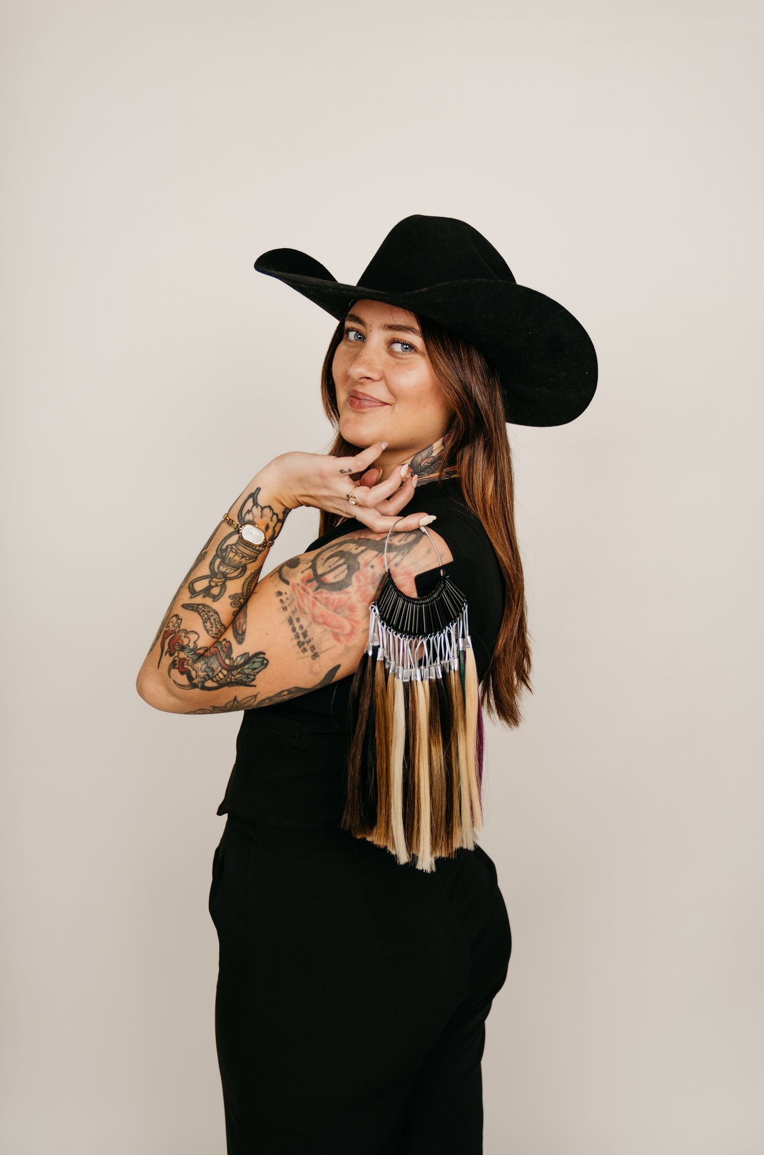 Tattooed woman in black cowboy hat and outfit with fringe necklace posing against light background.