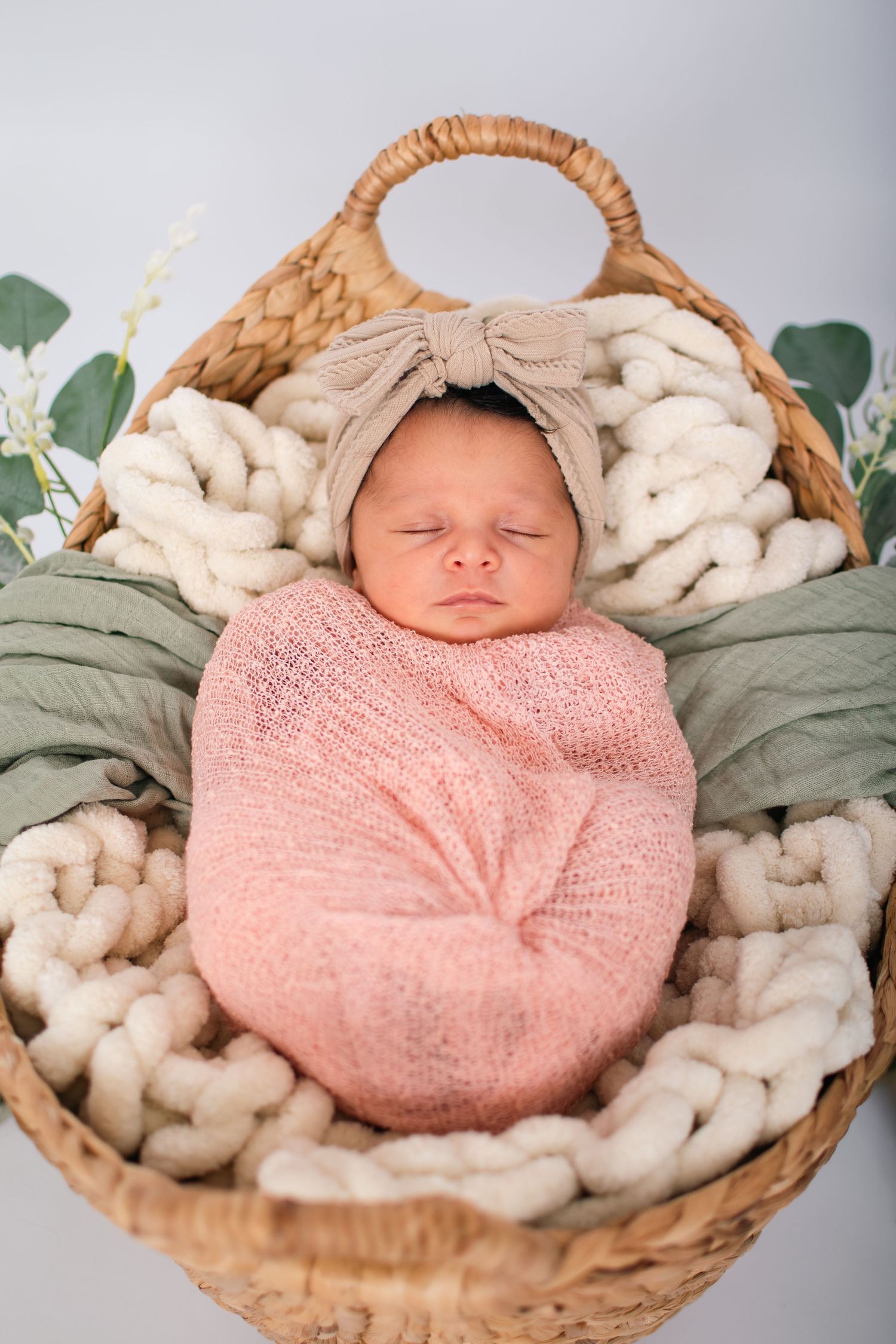 Sleeping newborn wrapped in pink swaddle with bow headband lying in woven basket on chunky knit blanket.