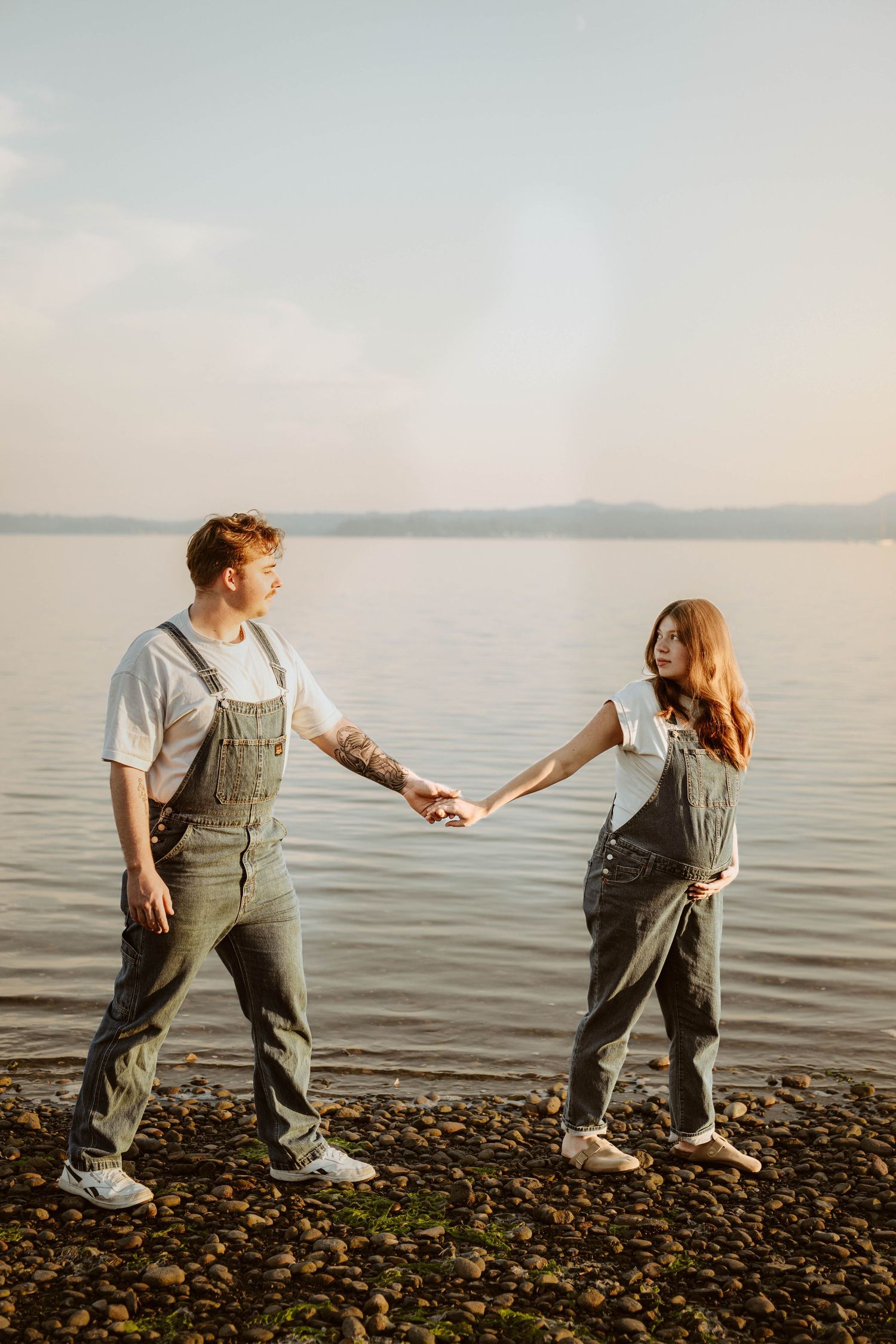 Couple walking hand in hand along rocky shoreline at sunset wearing matching overalls.