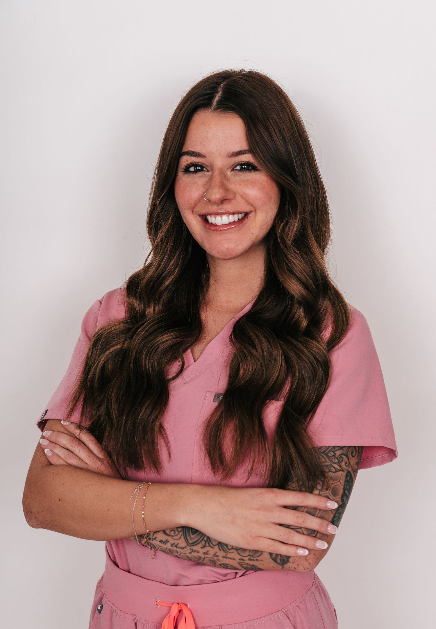 Smiling female healthcare professional in pink scrubs with tattoos and long brown hair, arms crossed.