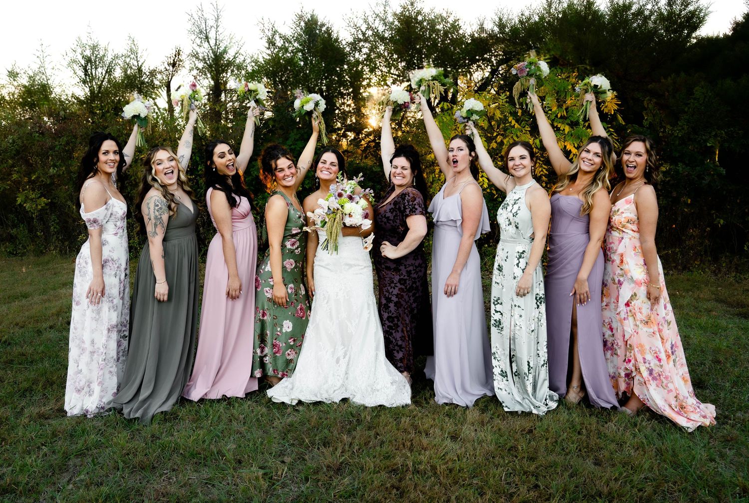 Wedding party group photo with bridesmaids in mix of purple and floral dresses outdoors.