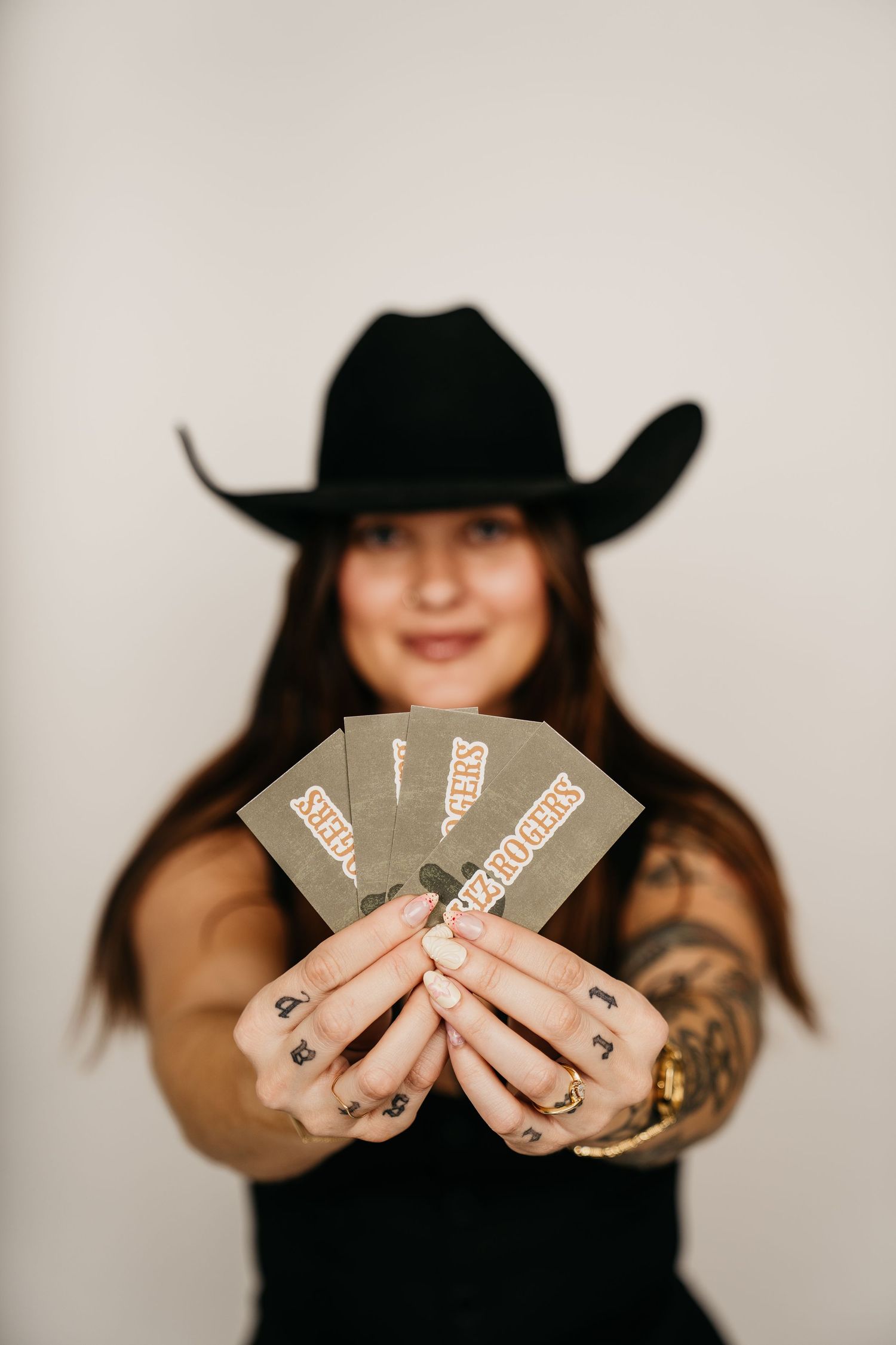 Tattooed woman in black cowboy hat holds fan of cards toward camera against white background.