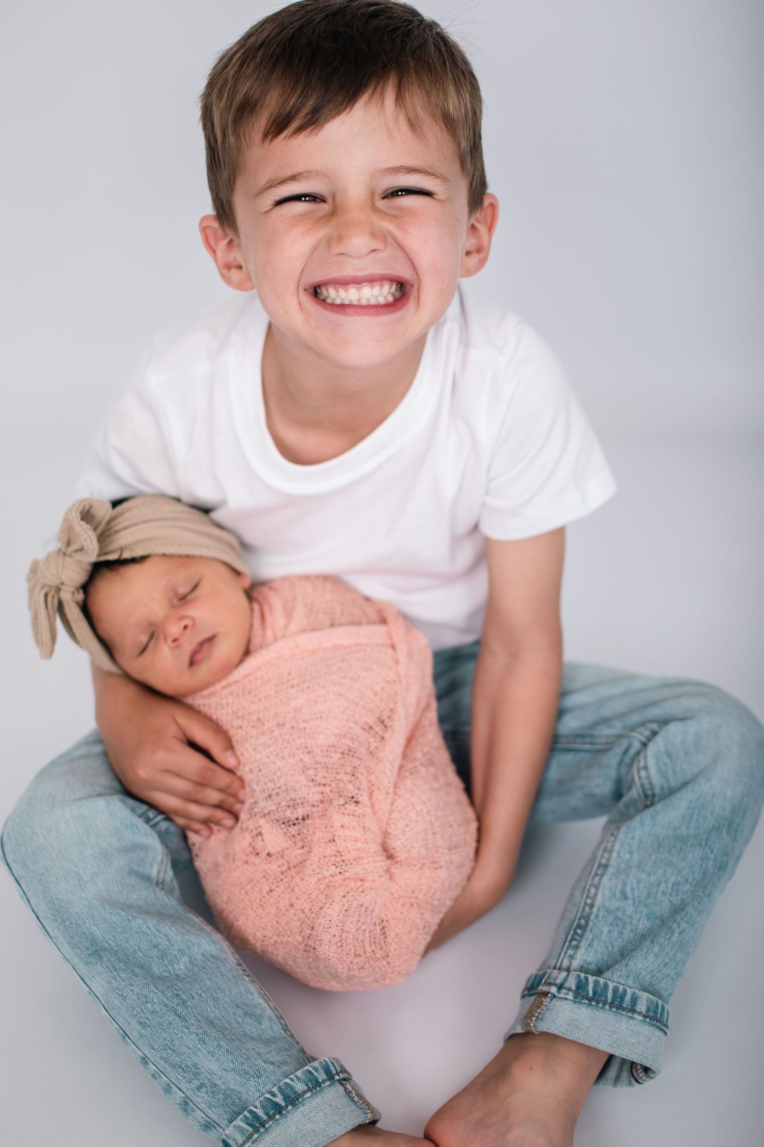 Studio portrait of young boy holding sleeping newborn wrapped in pink swaddle.