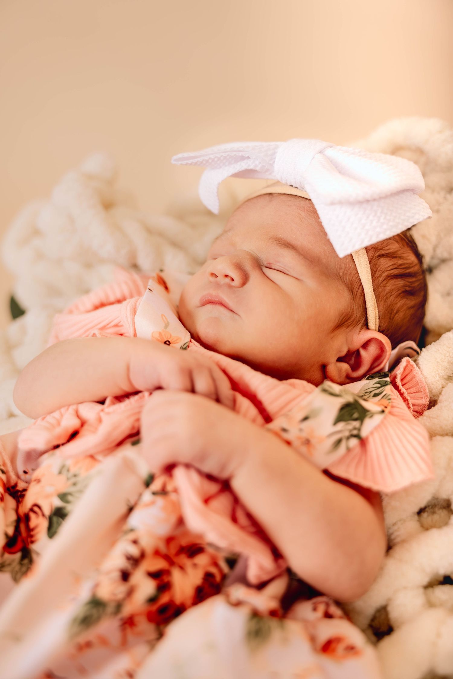 Sleeping newborn in floral outfit with white bow headband on soft blanket.