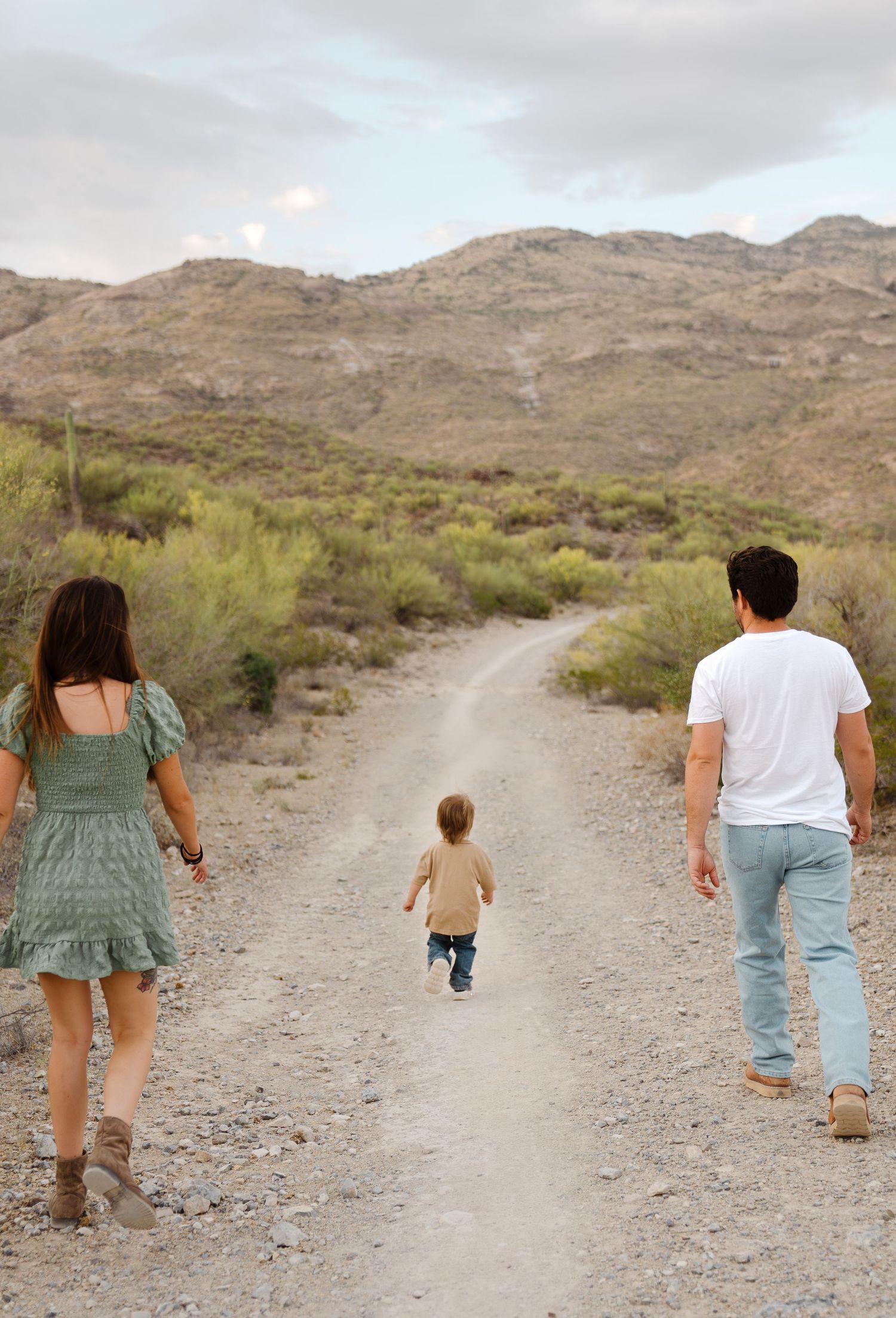 Family of three walking on desert trail, parents holding hands with toddler between mountains and shrubs.