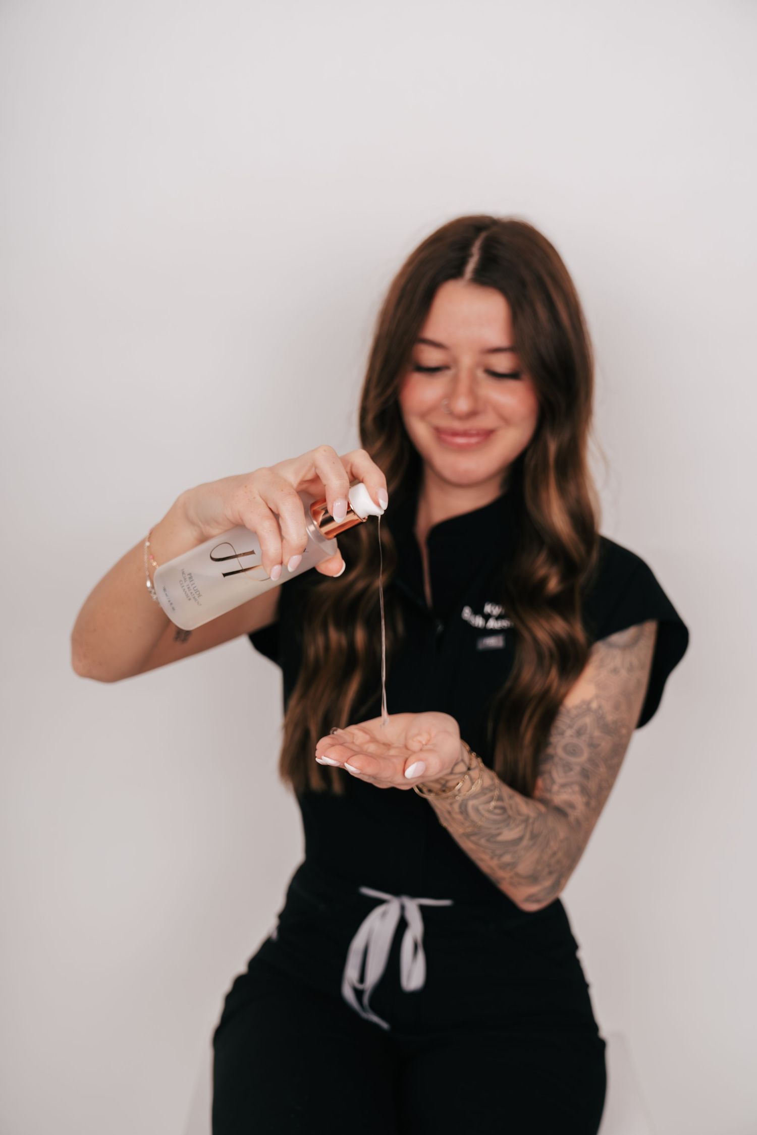 Woman in black outfit with tattoos dispensing clear liquid product into her hand against white background.