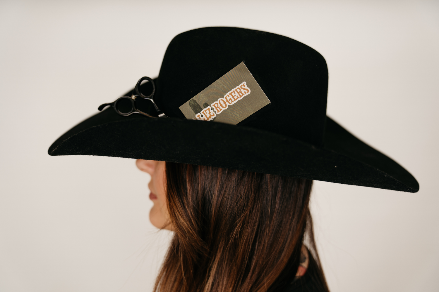 Woman wearing a wide-brim black felt hat with scissors and brand tag detail, side profile view.