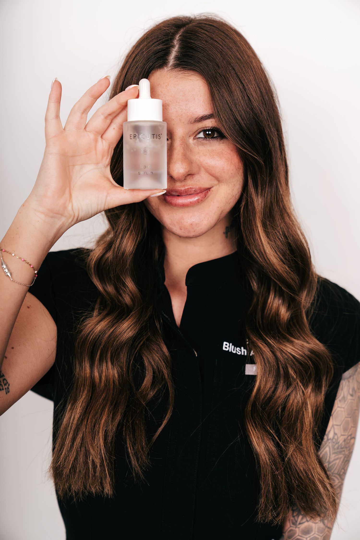 Woman in black Blush uniform holding a skincare serum bottle, showcasing beauty products with a smile.