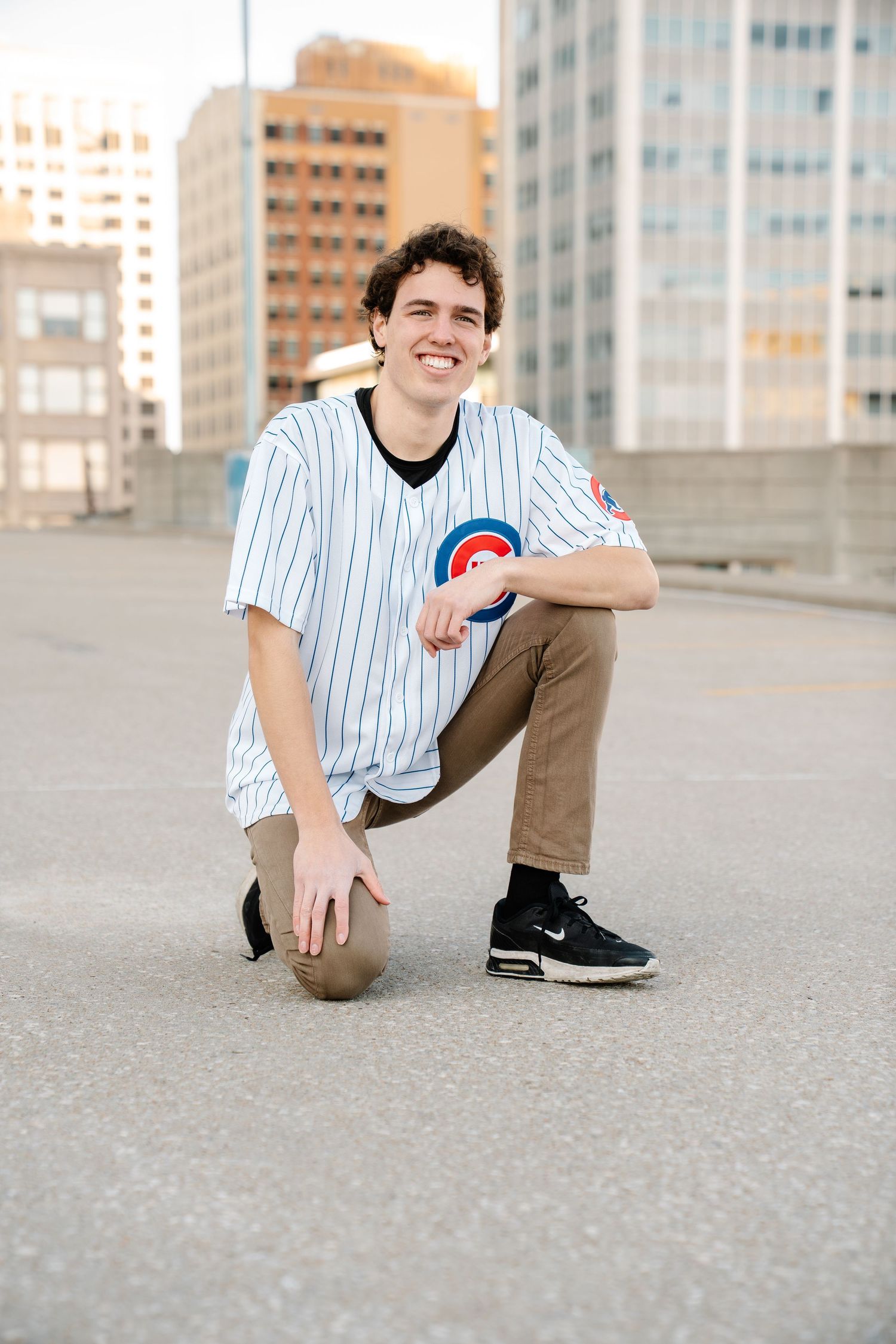 Smiling young man in Chicago Cubs jersey crouching on rooftop with city buildings in background.