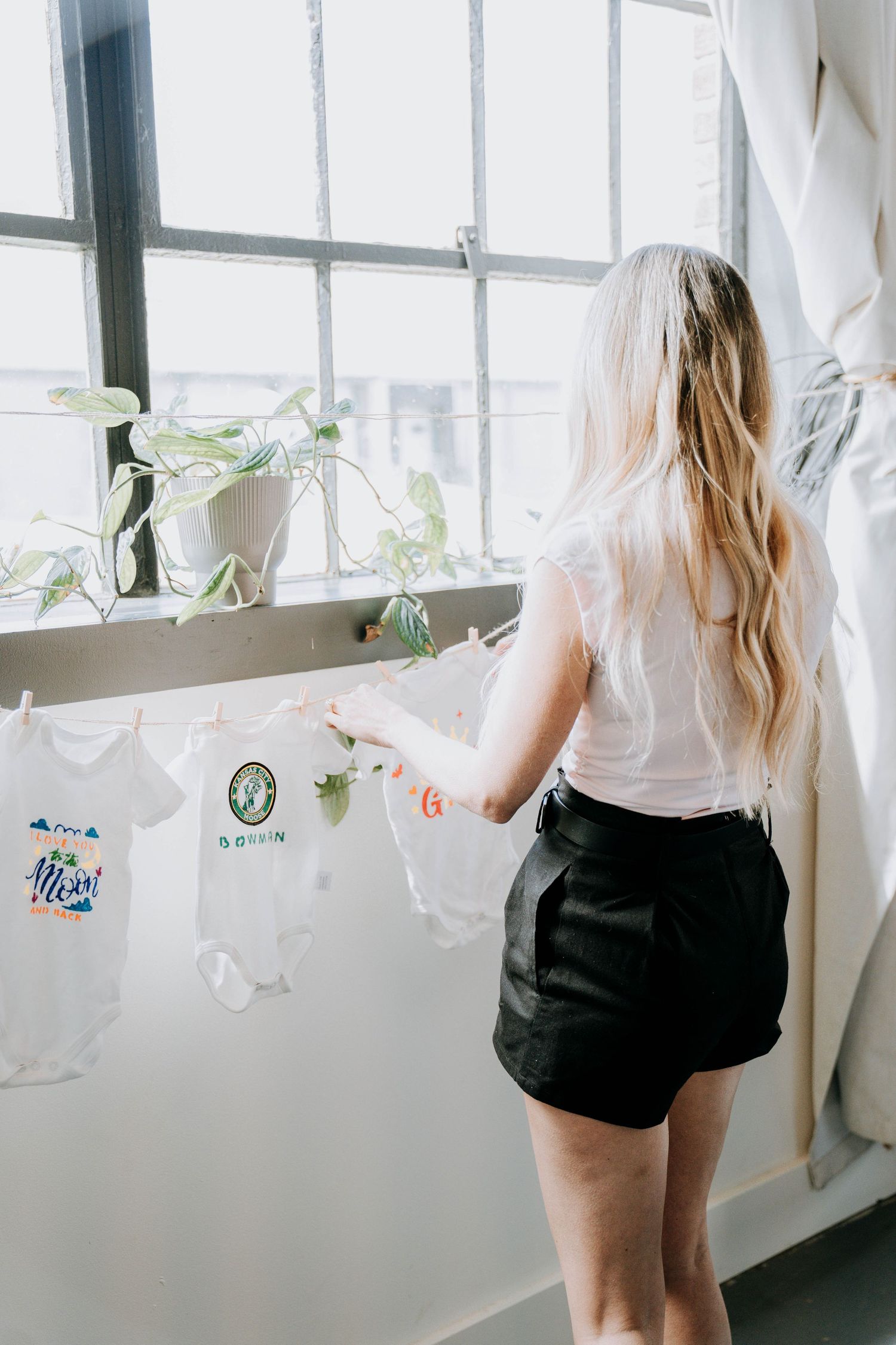 Woman in white shirt and black shorts looking out large windows with decorative hanging elements.